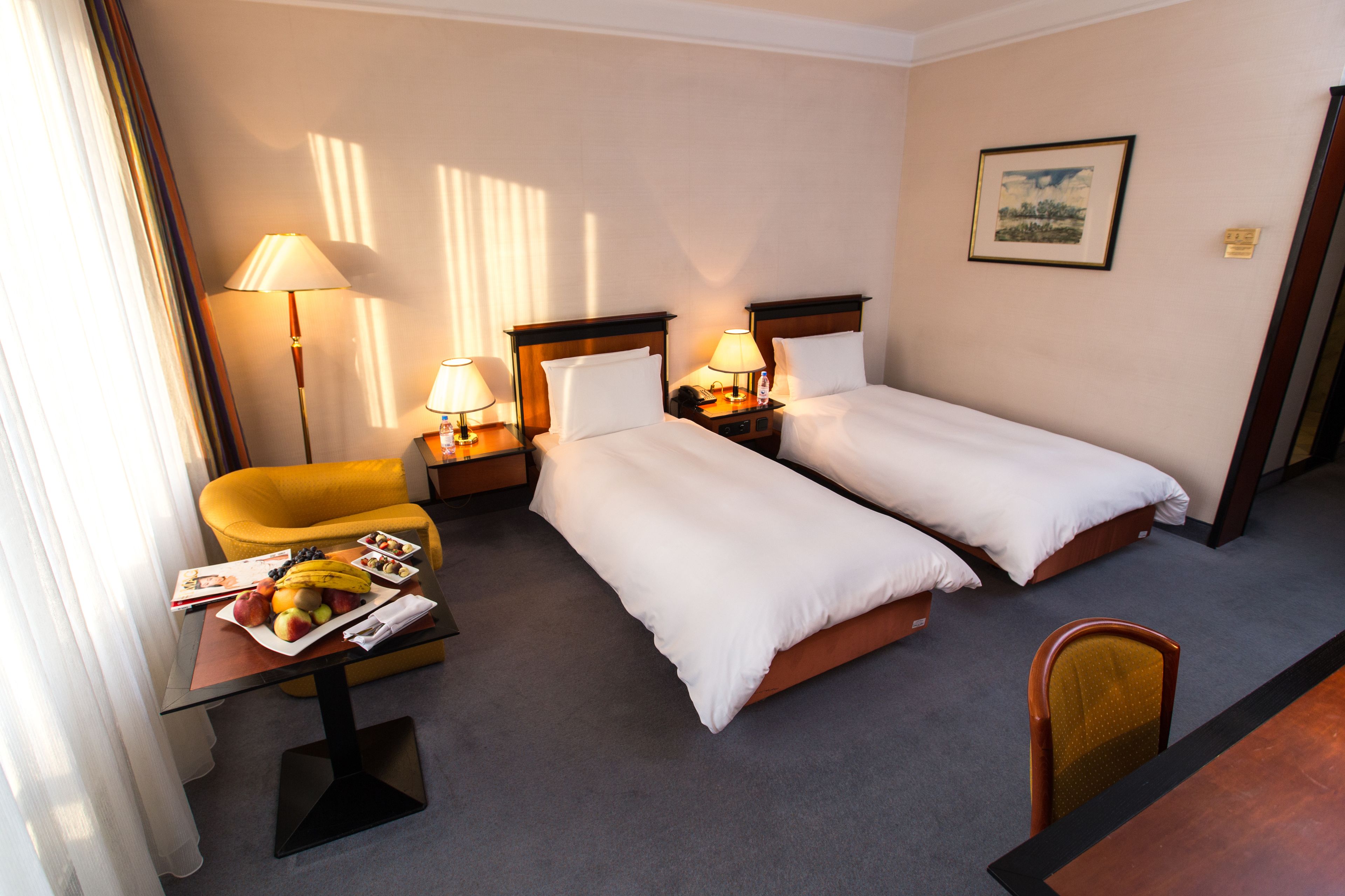 A hotel room with two single beds, a small table with a fruit bowl, a chair, and a lamp.