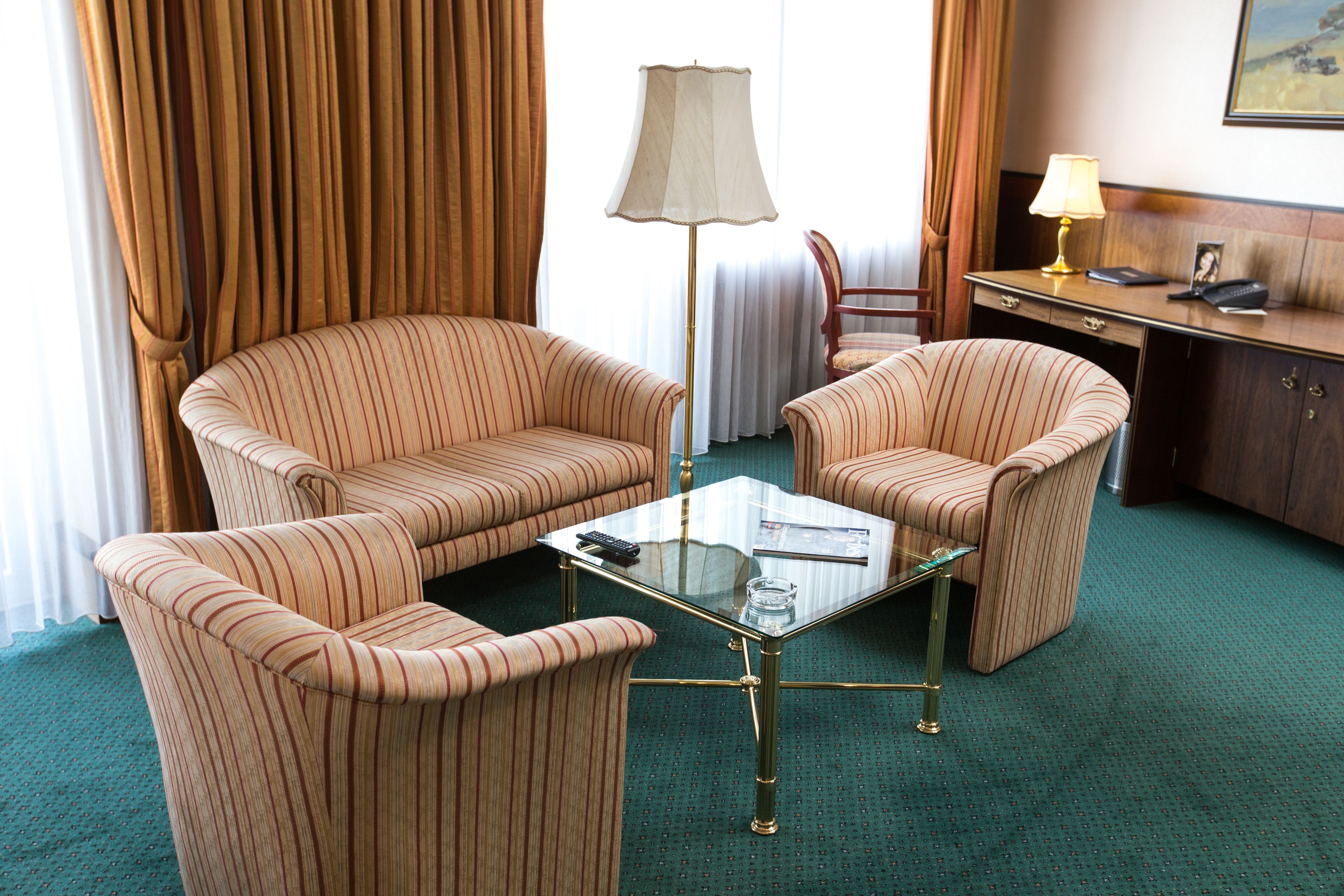Habitación de hotel con dos sillones a rayas, una mesa de centro de vidrio, un escritorio con lámpara y vistas al exterior a través de cortinas.