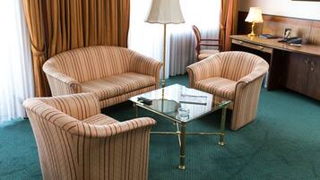 Habitación de hotel con dos sillones a rayas, una mesa de centro de vidrio, un escritorio con lámpara y vistas al exterior a través de cortinas.