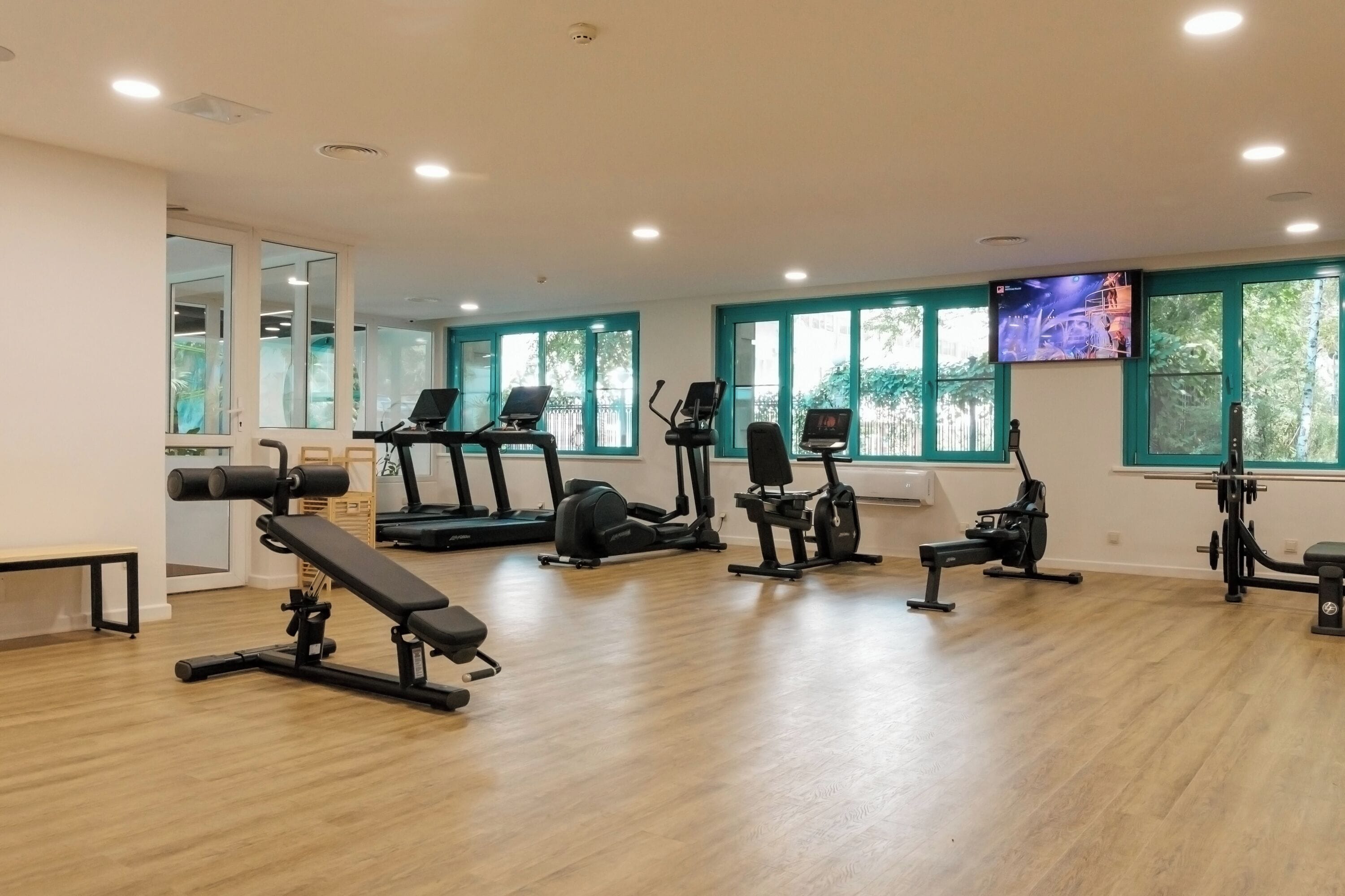 Un gimnasio con diversos equipos de ejercicio, ventanas amplias y una pantalla de televisión.
