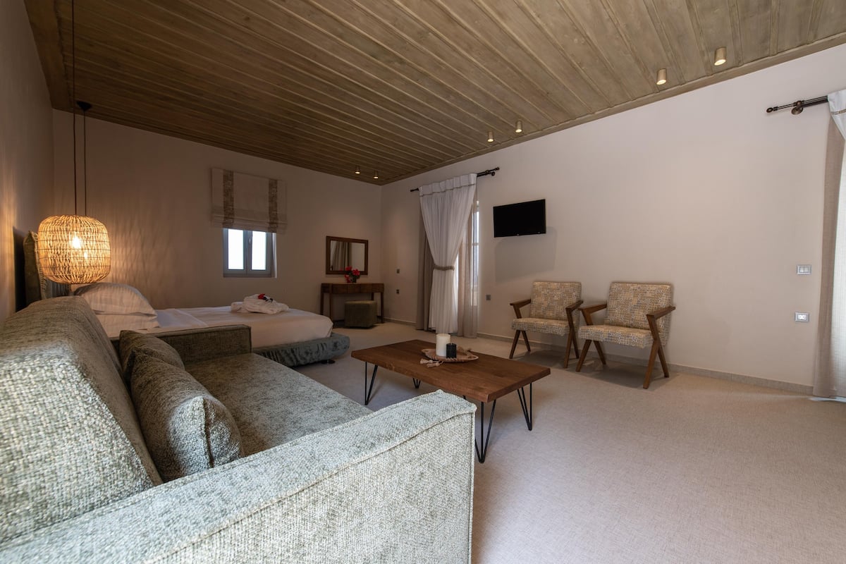 A spacious room with a wooden ceiling, a large sofa, two armchairs, a small table with a candle, a TV mounted on the wall, and a window with curtains.
