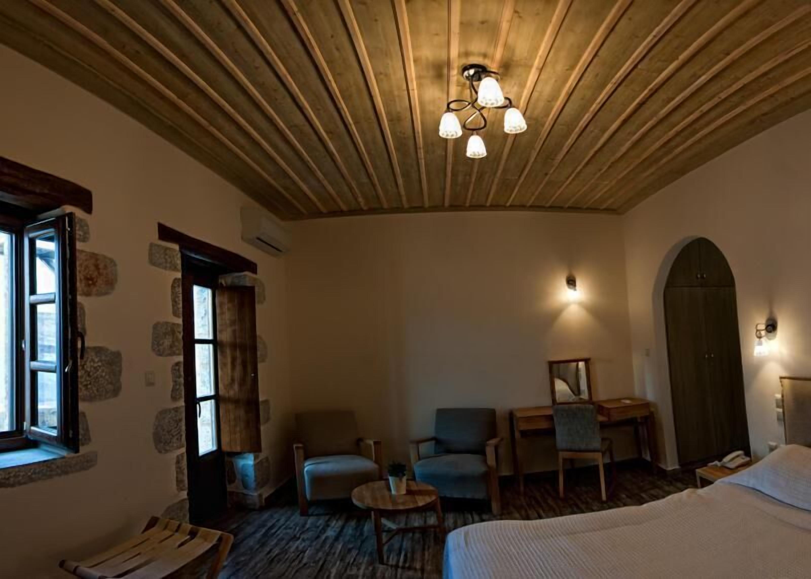 A cozy room with a wooden ceiling, stone walls, a bed, two armchairs, a small table, and a desk with a chair.