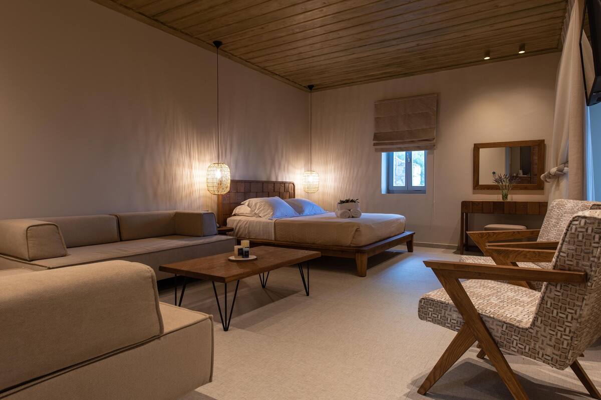 A modern bedroom with a bed, sofa, and armchair, wooden ceiling, and a window with a view.