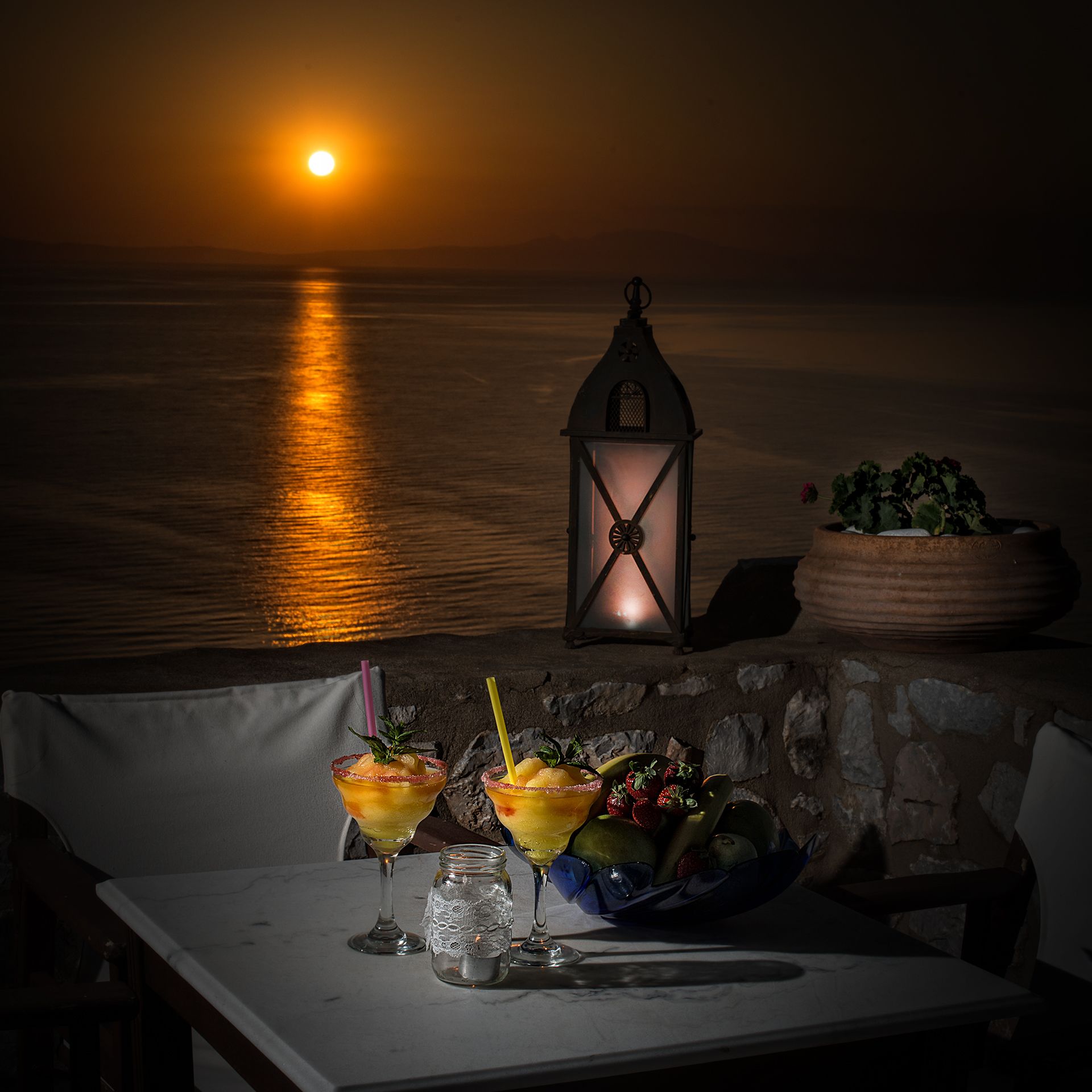 Uma mesa posta para dois, com bebidas, frutas, um candeeiro e vista para o oceano ao pôr do sol.