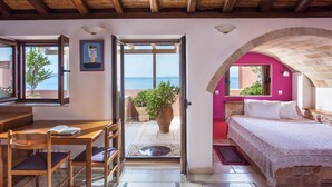 A room with a wooden ceiling, a bed with a stone arch, a desk with chairs, and a view of the sea through a glass door. - Aktaion Hotel (Monemvasia)