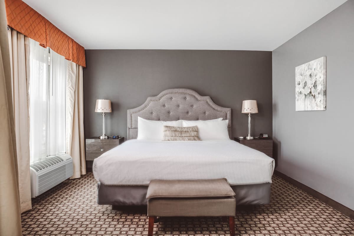 A hotel room with a large bed, two bedside lamps, a bedside table, a window with curtains, a radiator, a patterned floor, and a piece of wall art.