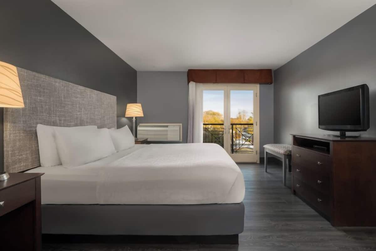 A modern hotel room with a large bed, bedside lamps, a television, a dresser, and a balcony with a view.