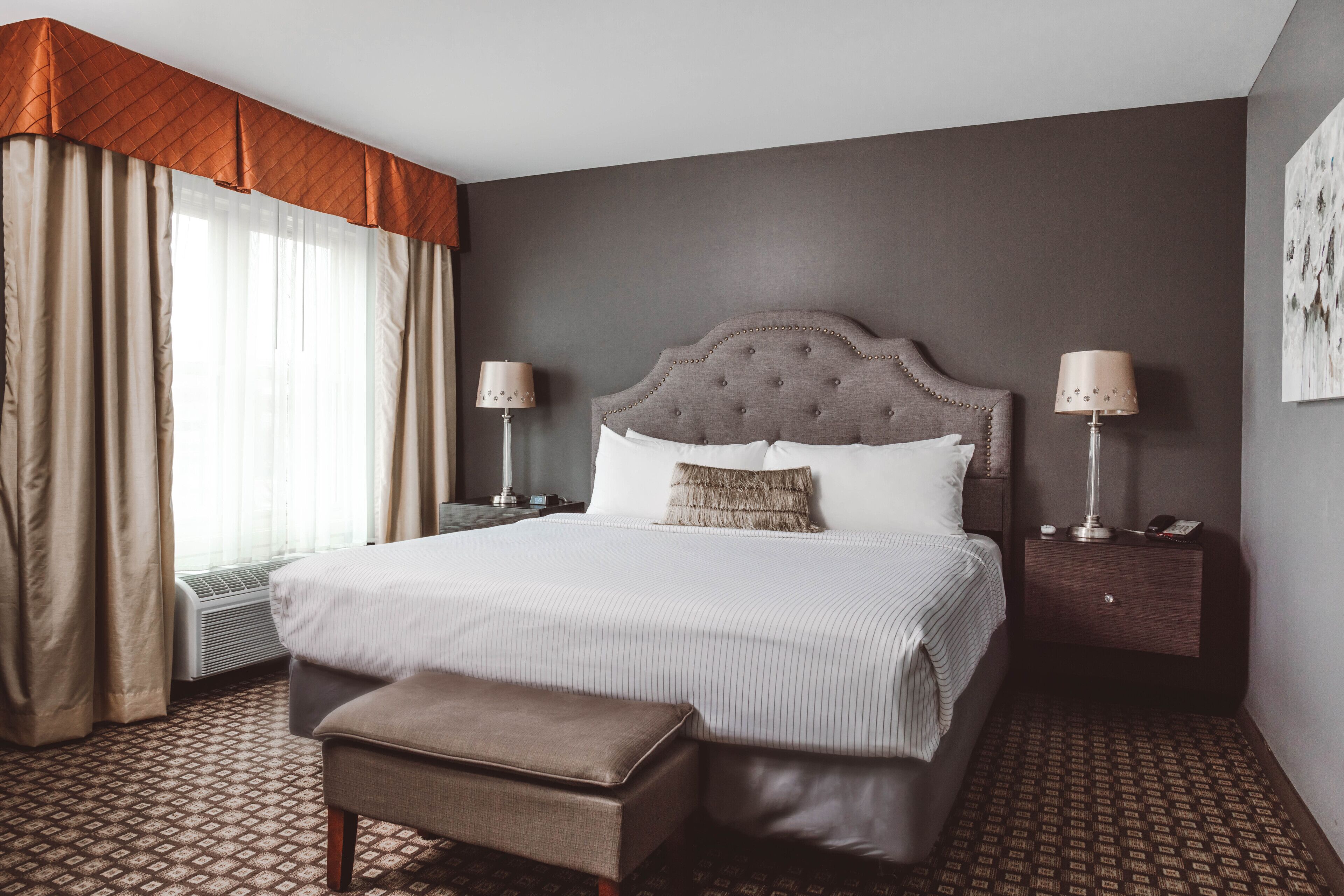 A hotel room with a large bed, two bedside tables with lamps, a bench, a patterned carpet, and a window with curtains.