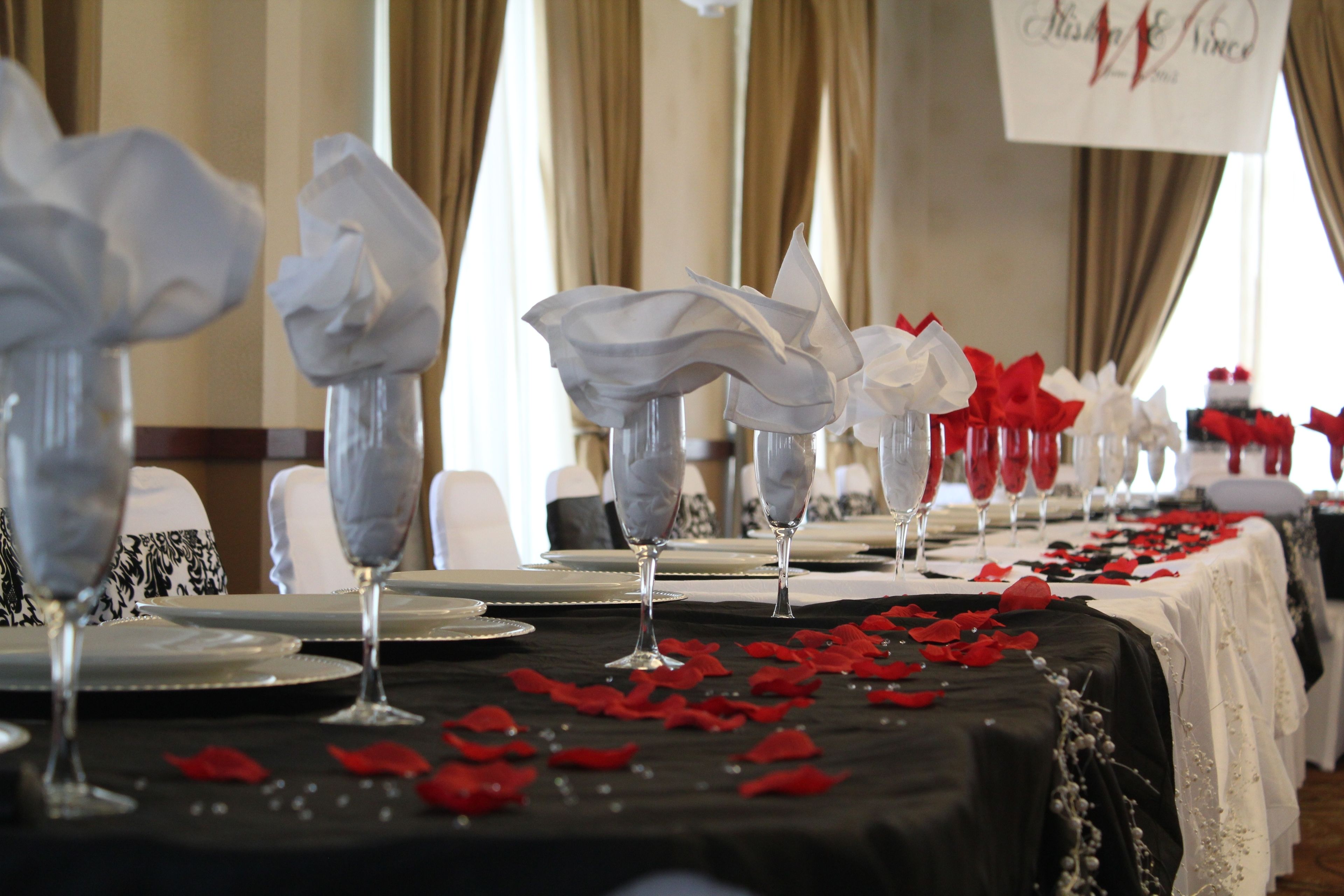 A banquet table with white and red rose petals, clear glassware, and a black tablecloth.