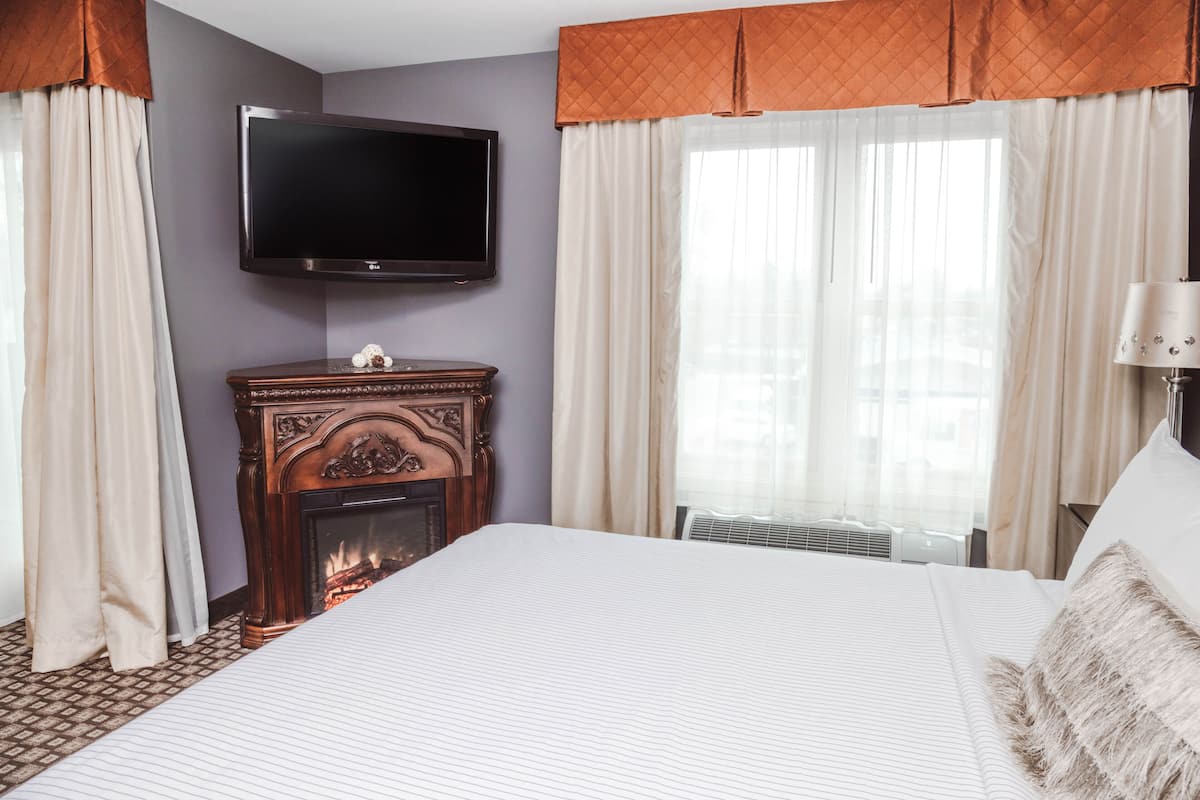 A hotel room with a large bed, a flat-screen TV mounted on the wall, a fireplace, and a window with sheer curtains.