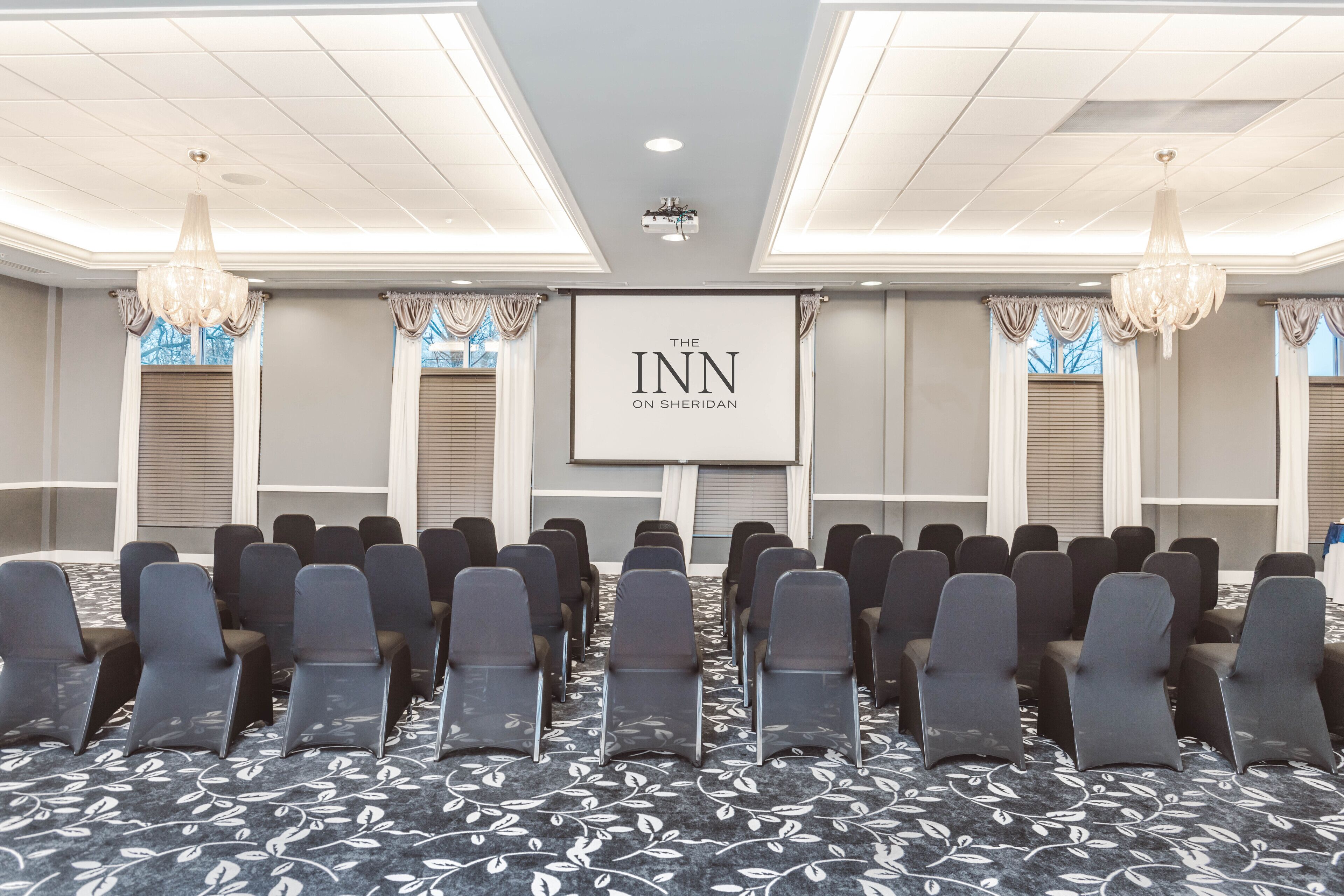 A conference room with rows of chairs, a projector screen displaying "The Inn on Sheridan," and chandeliers.