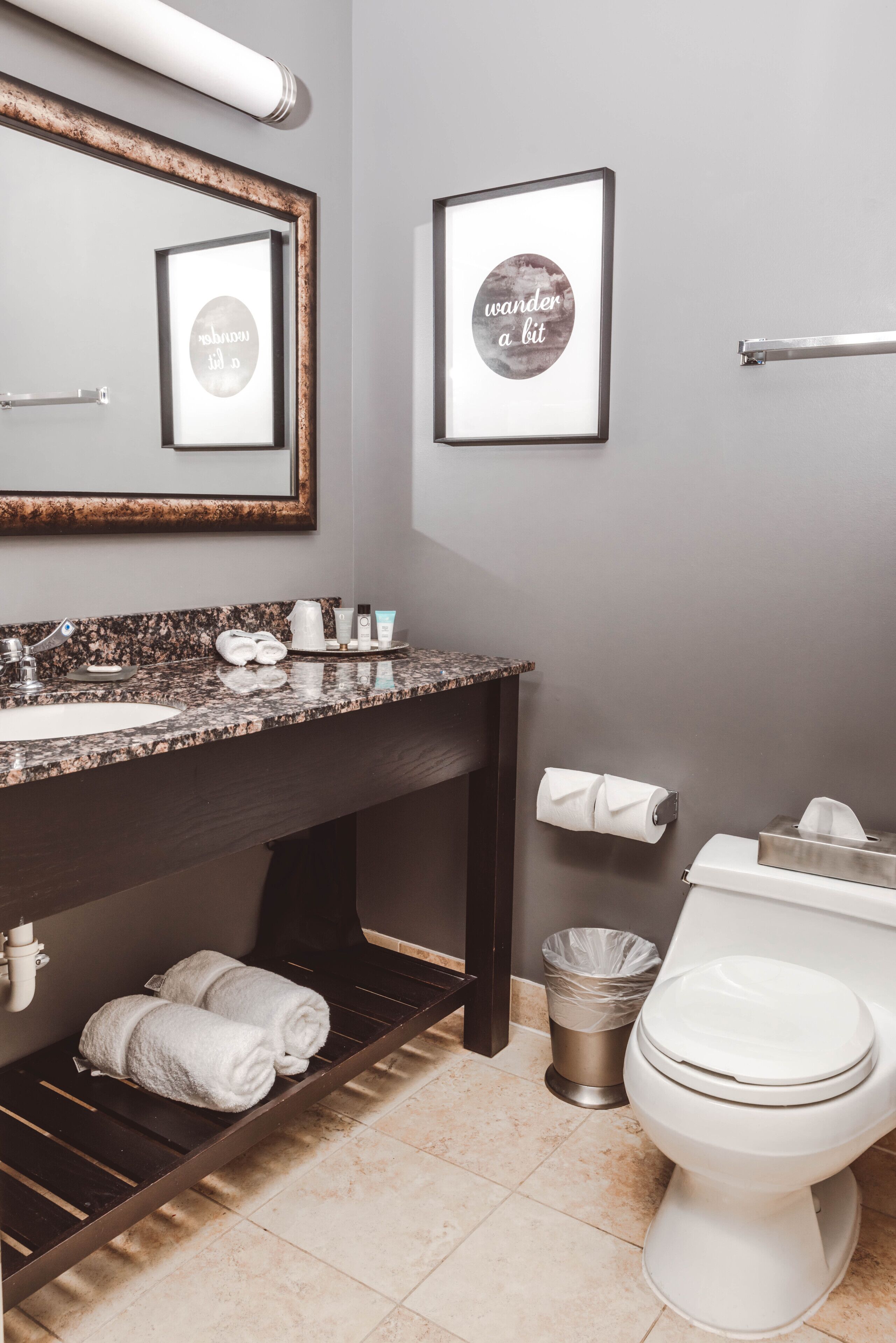 A bathroom with a granite countertop, a mirror, two framed pictures, a towel rack, a toilet, and a trash can.