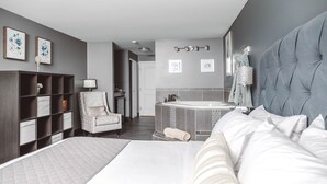 A modern hotel room with a large bed, a seating area with a chair, a small table, a vanity with a mirror, and a bathtub.