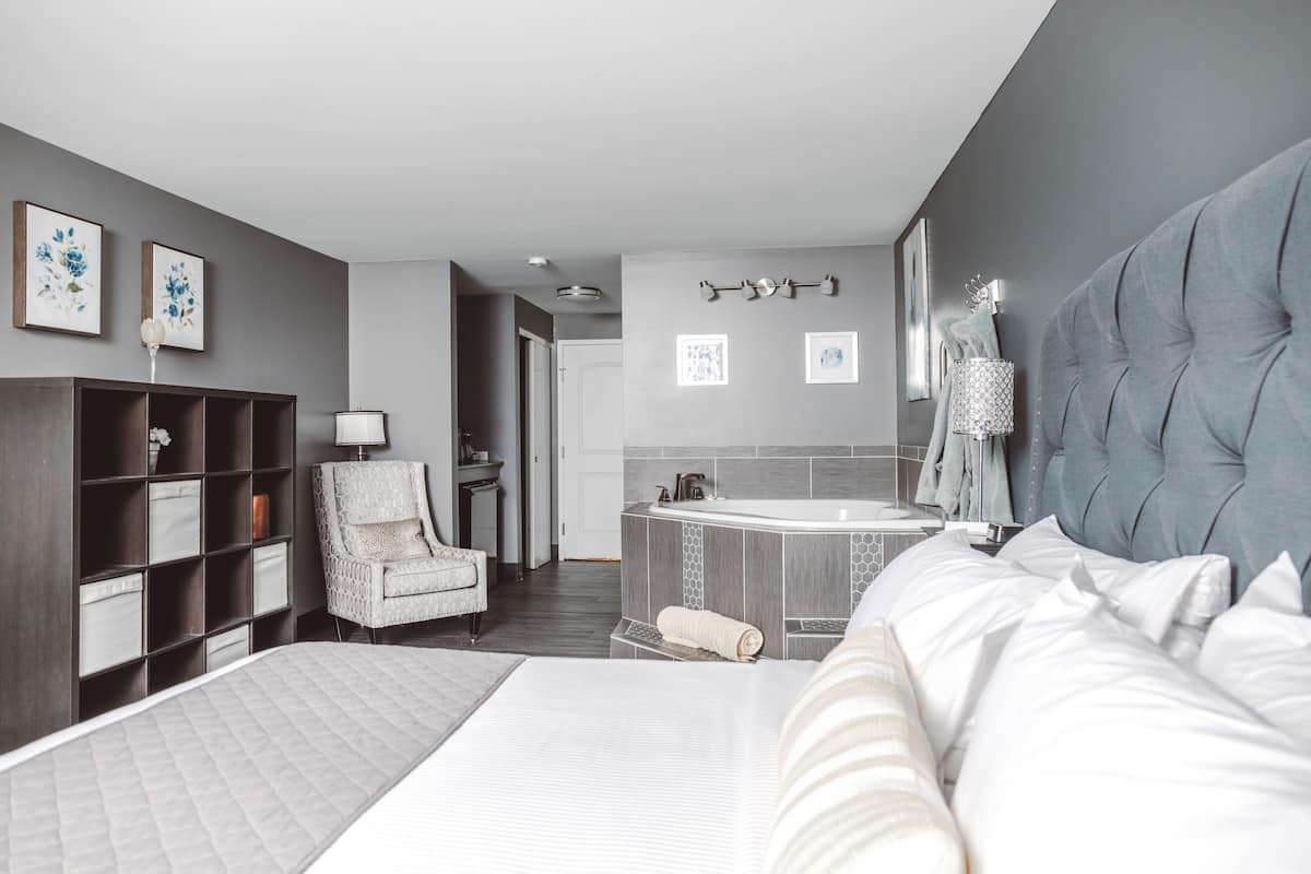 A modern hotel room with a large bed, a seating area with a chair, a small table, a vanity with a mirror, and a bathtub.