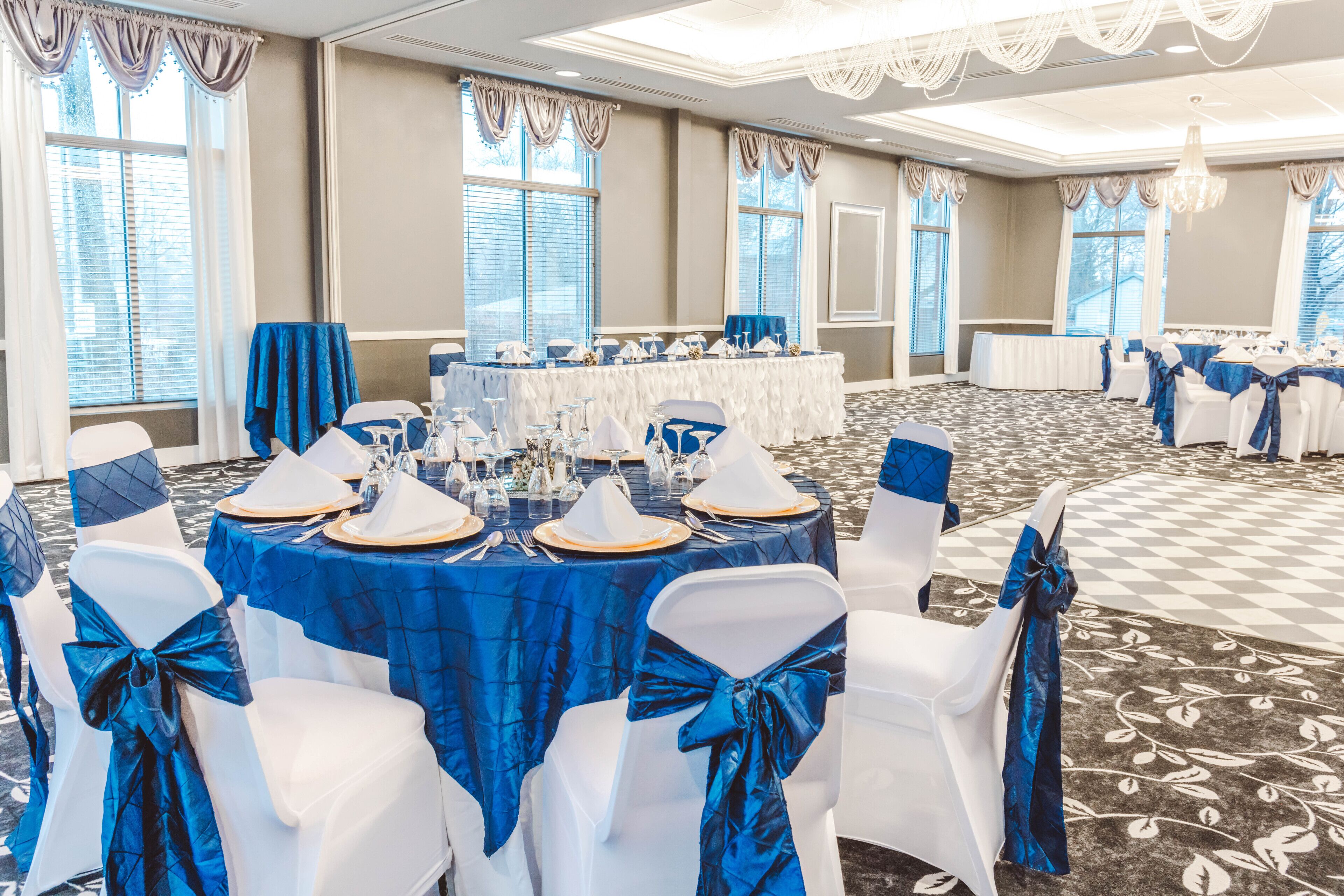 A banquet hall with round tables set for a formal event, featuring white chairs with blue sashes and a checkered carpet.