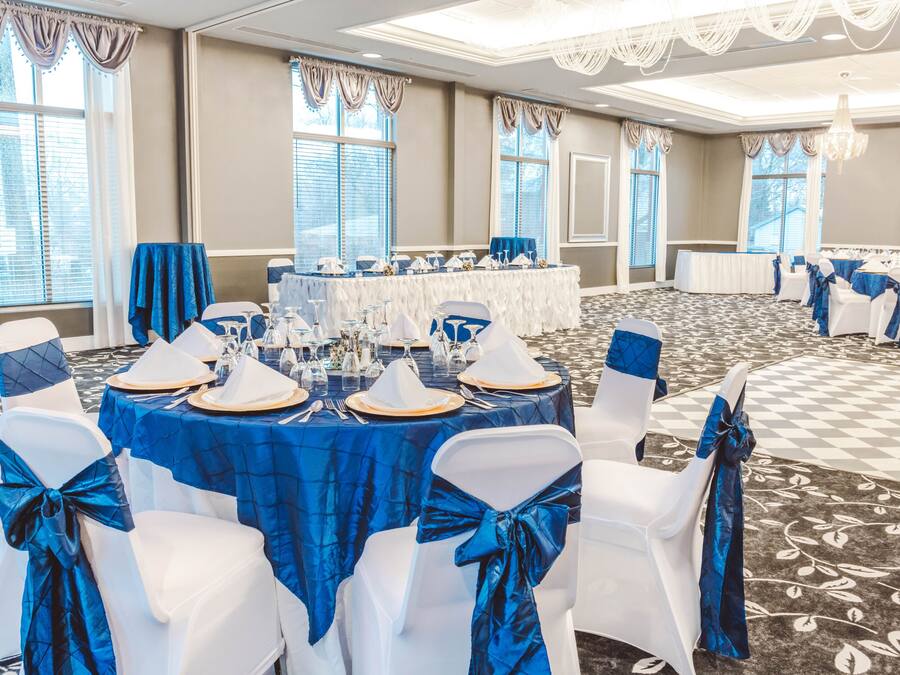 A banquet hall with round tables set for a formal event, featuring white chairs with blue sashes and a checkered carpet.