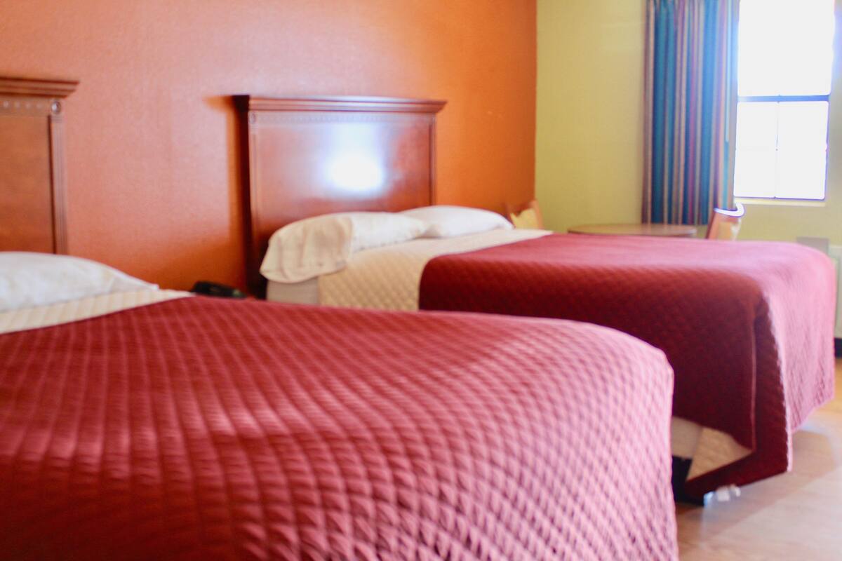 Quarto de hotel com duas camas, cabeceira de madeira, janela com cortinas e uma mesinha.