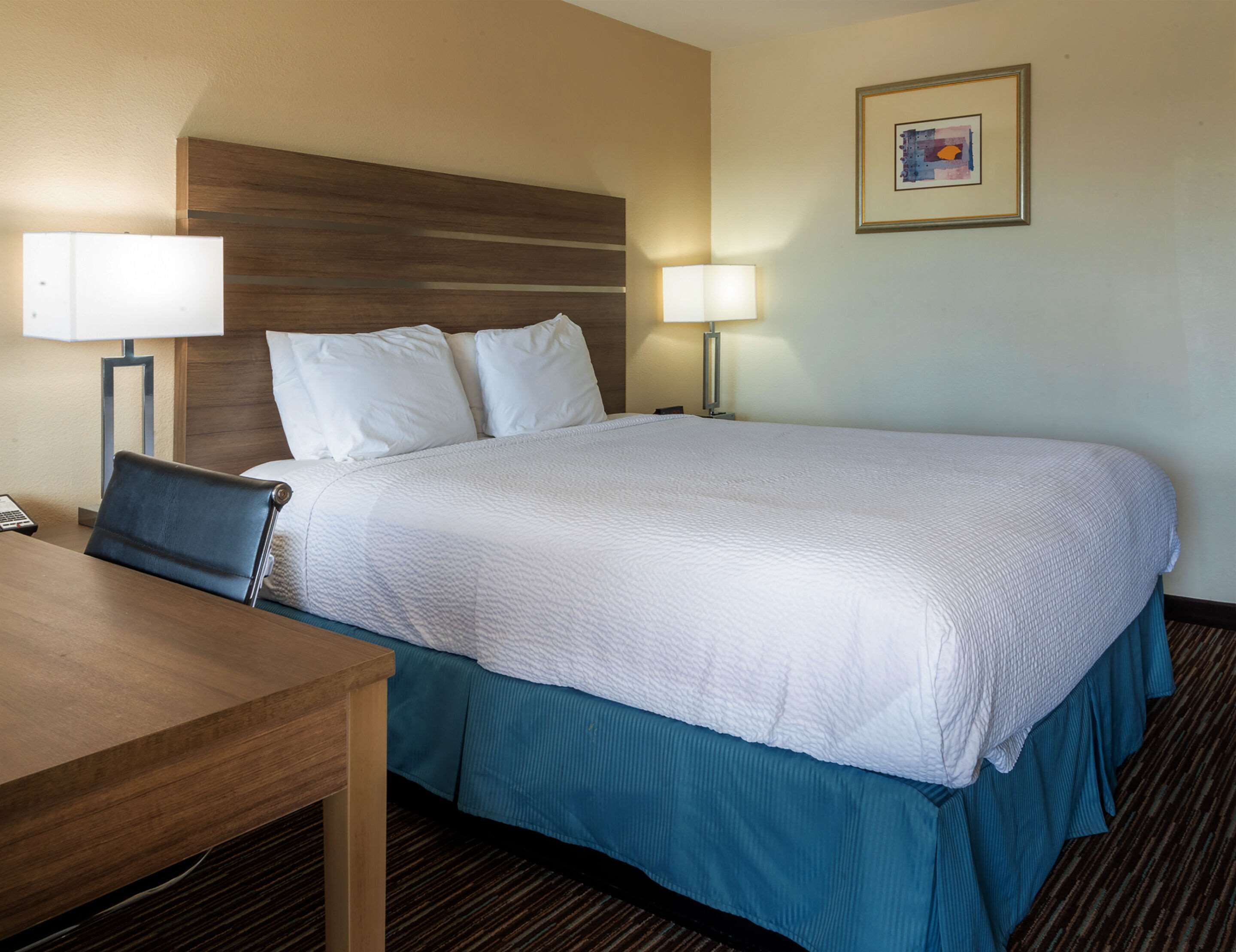 Una habitación de hotel con una cama grande, dos lámparas de noche, un cabecero de madera, un escritorio con silla y un cuadro en la pared.