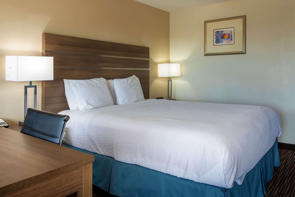 A hotel room with a large bed, two bedside lamps, a wooden headboard, a desk with a chair, and a framed picture on the wall.