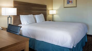 A hotel room with a large bed, two bedside lamps, a wooden headboard, a desk with a chair, and a framed picture on the wall.