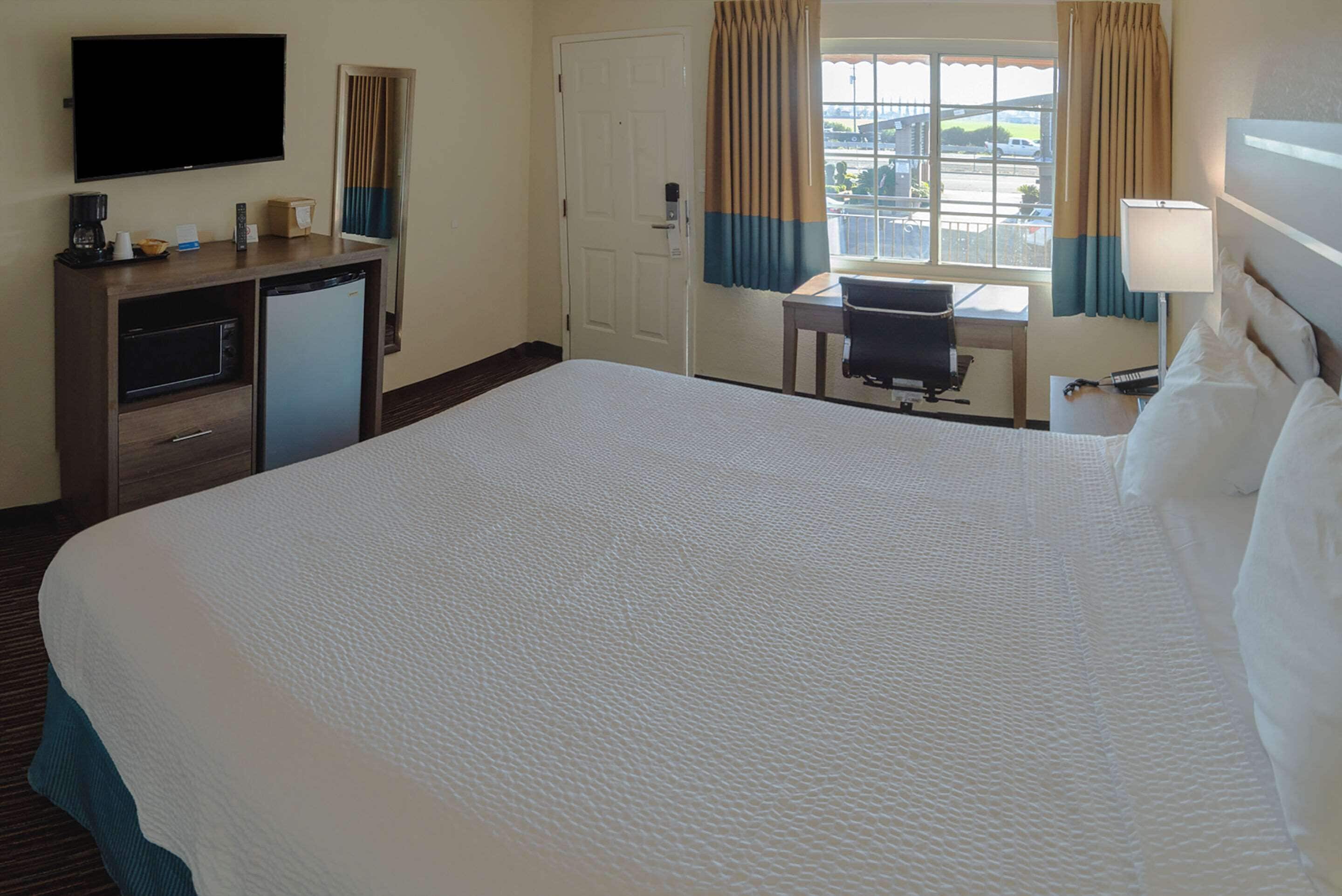 Habitación de hotel con una cama grande, un escritorio con silla, televisión, cafetera y ventana con cortinas.