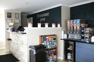 A reception area with a counter, a bookshelf with magazines, a refrigerator with beverages, and a wall with the hotel name.