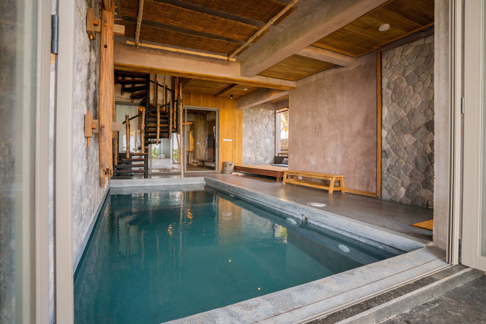 Piscina con pared de piedra y vigas de madera, una banca y una escalera que lleva a otra habitación.