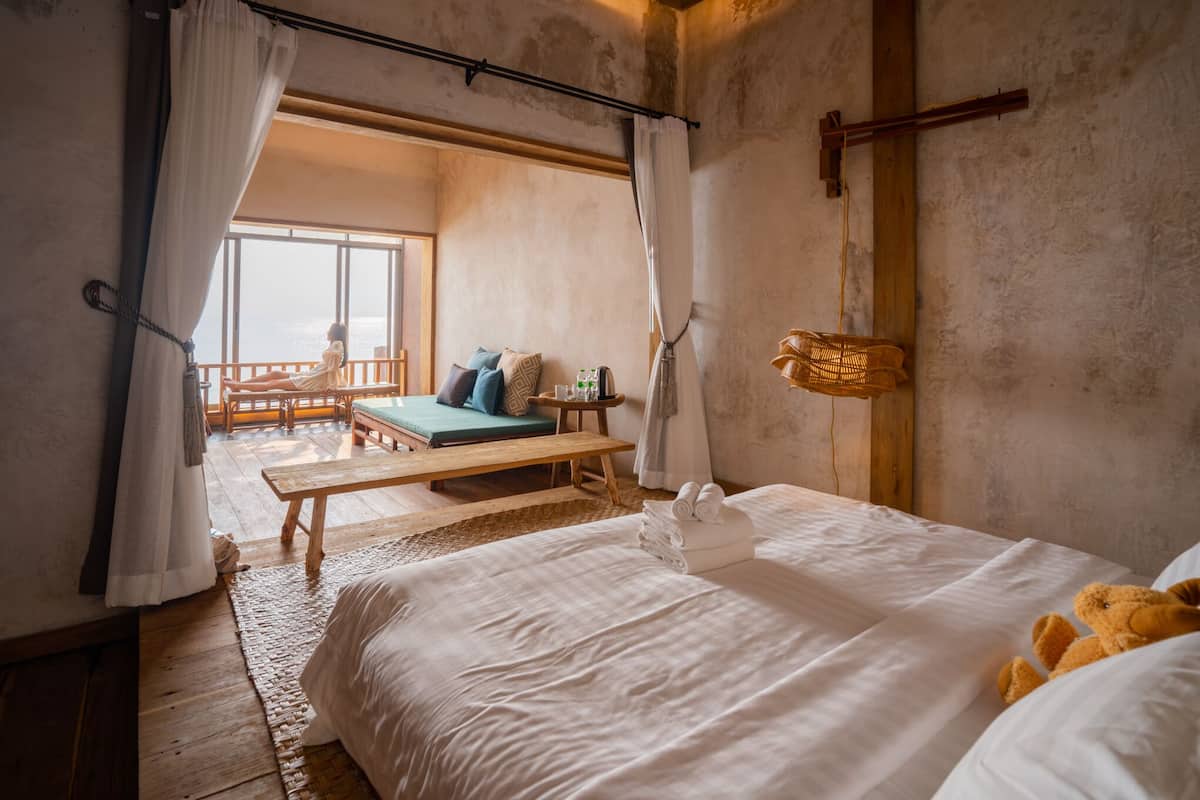 Een slaapkamer met een groot bed, een houten bankje en uitzicht op zee.