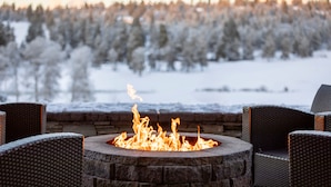 A stone fire pit with flames, surrounded by wicker chairs, overlooking a snowy landscape with trees. - Running Y Resort (Klamath Falls)