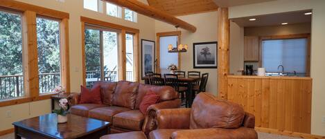 A spacious living area with a large L-shaped leather sofa, a coffee table, and a dining table with chairs. There are large windows providing natural light and a view of the outdoors.