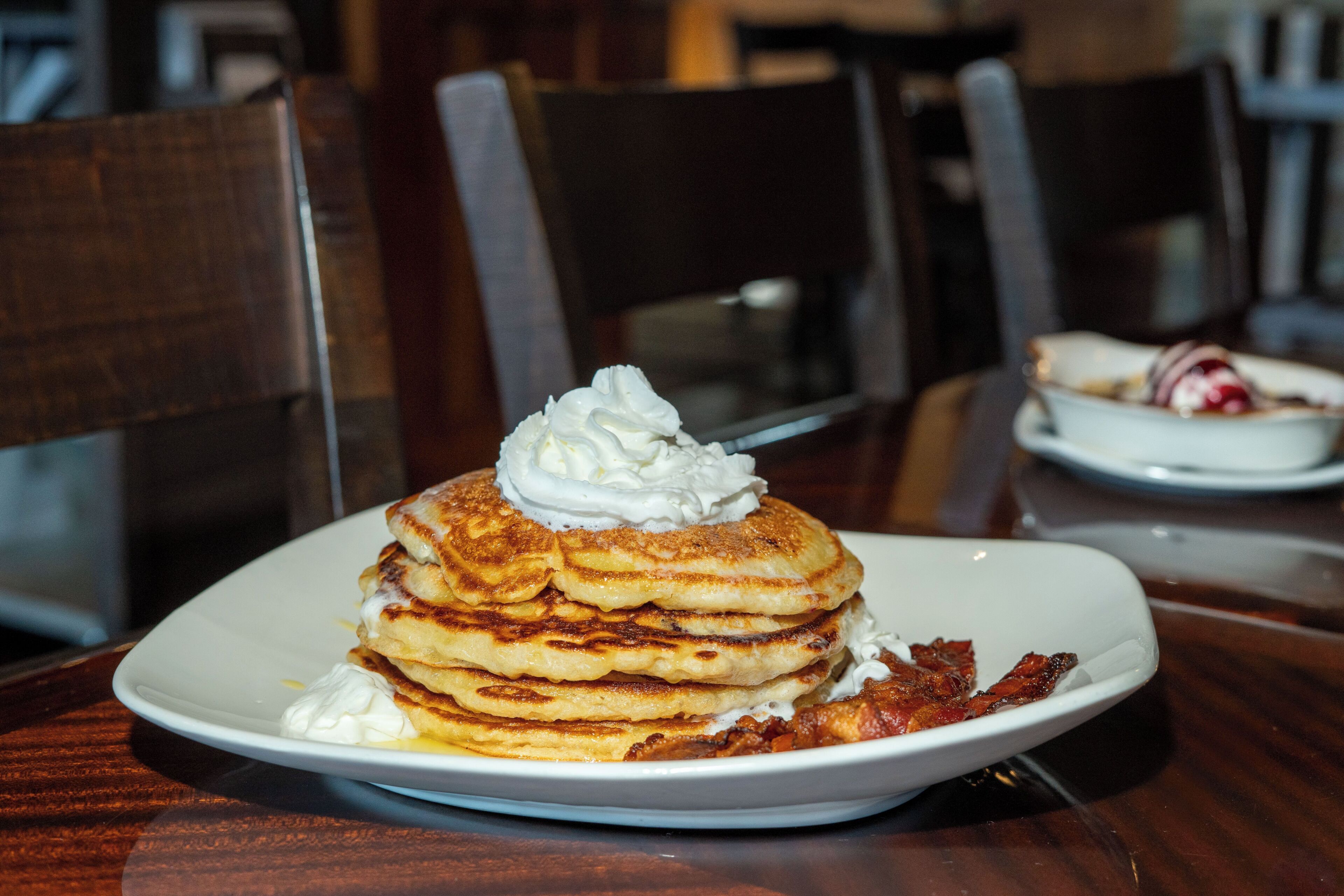A stack of pancakes with whipped cream on a white plate on a wooden table.