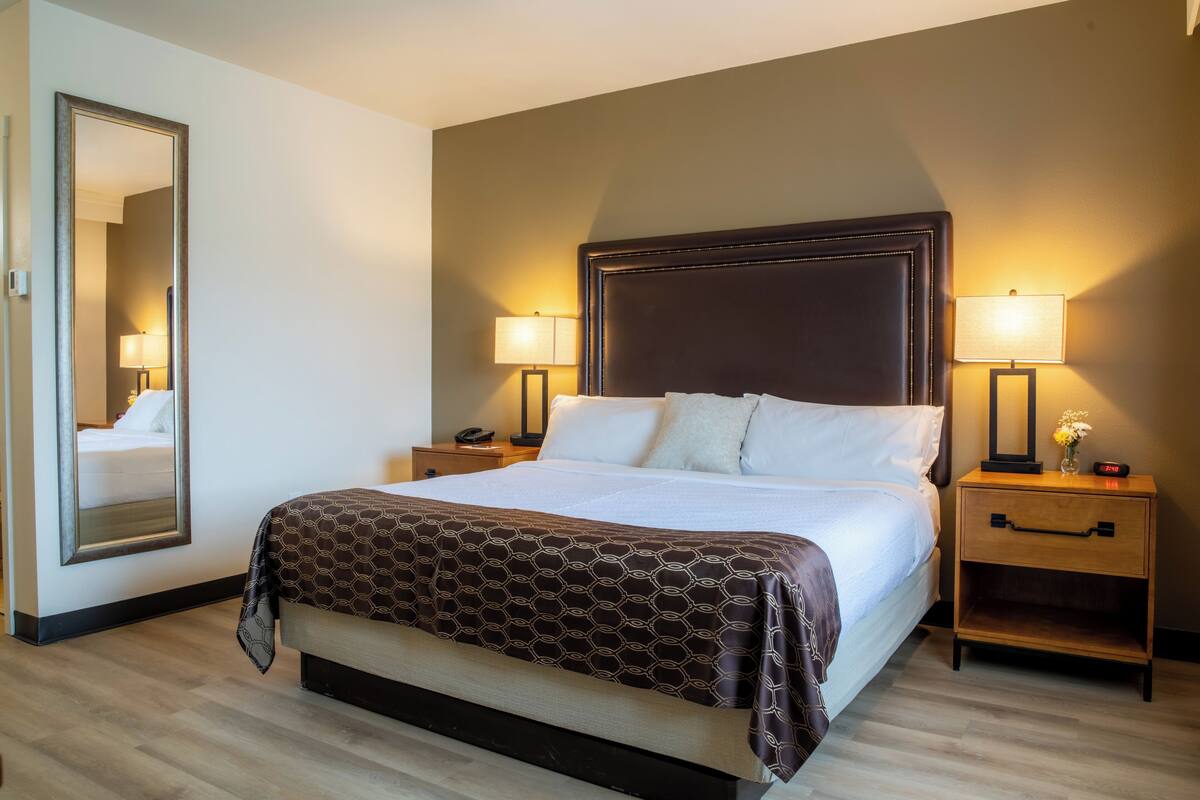 A hotel room with a large bed, two bedside tables with lamps, a nightstand, a mirror, and a wooden floor.