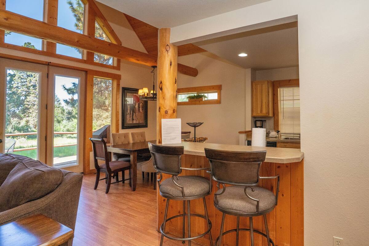 A spacious living area with a large window, a dining table with chairs, a bar area, and a kitchen visible through an open doorway.