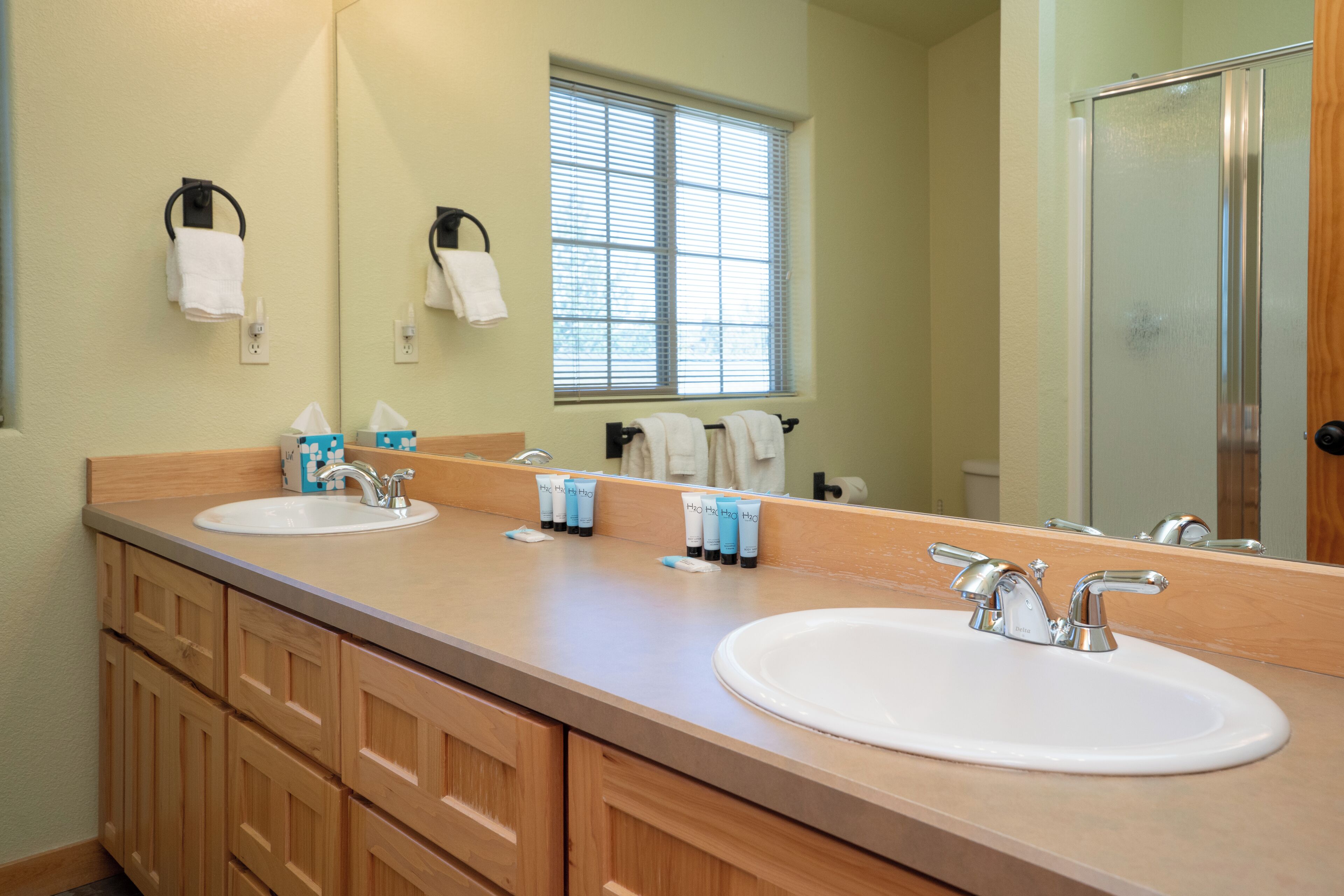 A bathroom with two sinks, wooden cabinets, a large mirror, and a window with blinds.
