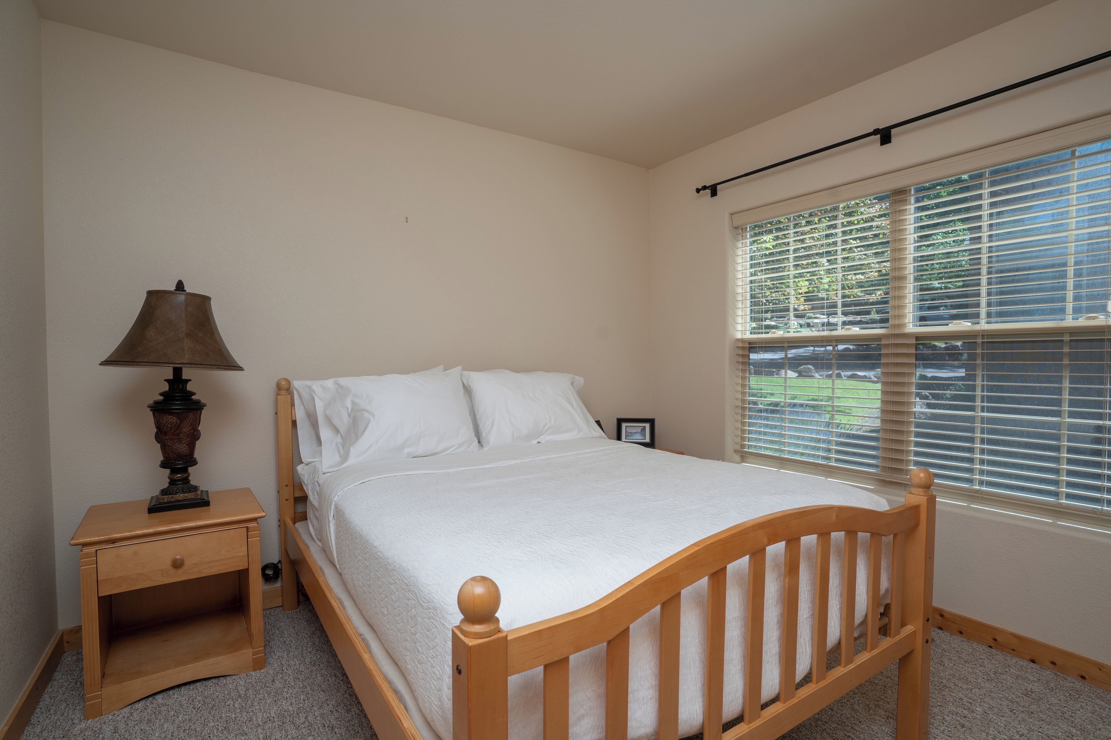 A bedroom with a wooden bed, a nightstand, a lamp, and a window with blinds.