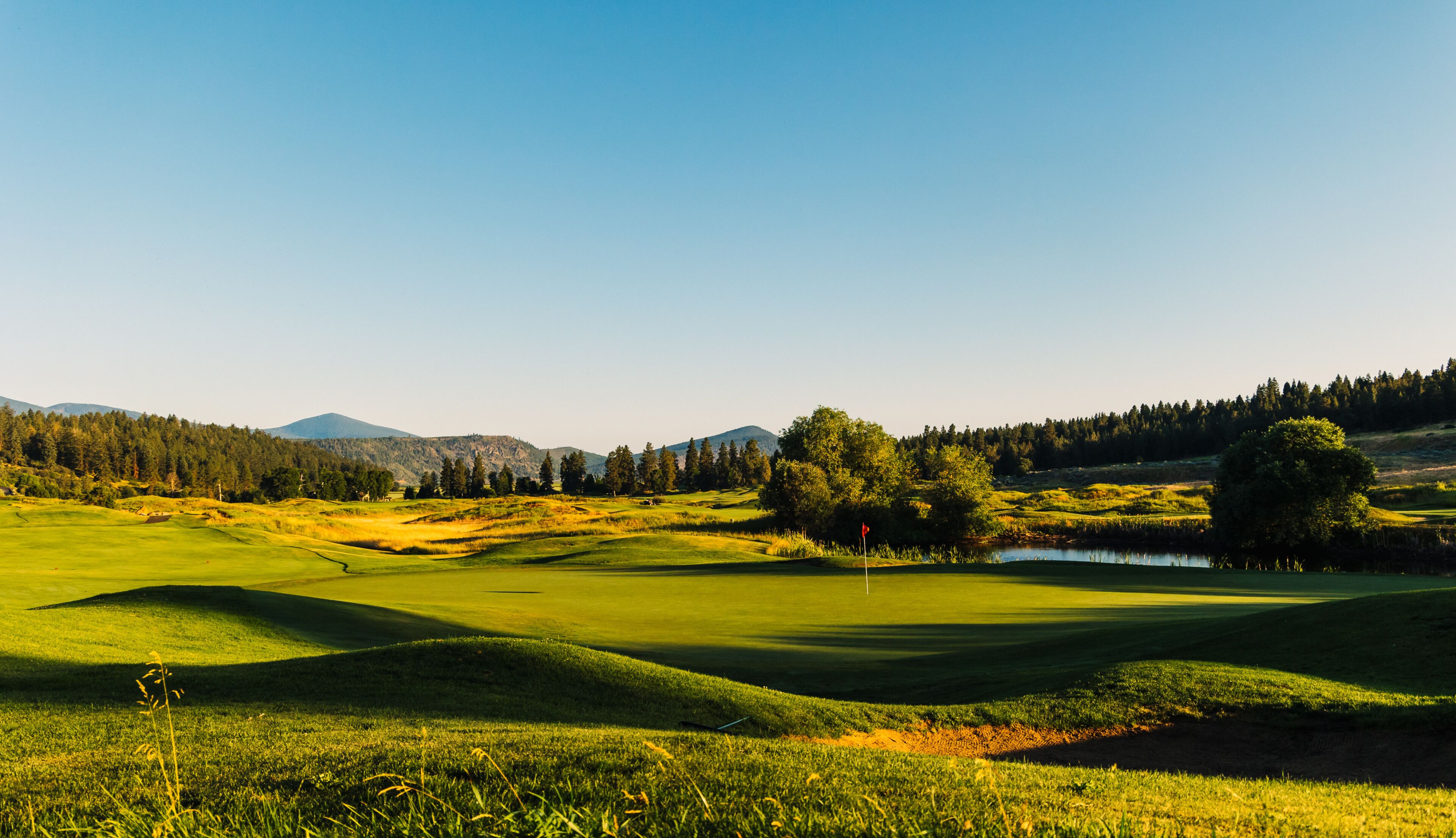 Un parcours de golf avec des collines douces, un étang et un mât de drapeau.