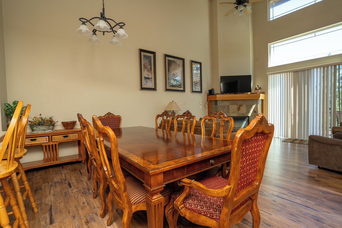 A dining area with a wooden table and chairs, a chandelier, framed pictures on the wall, and a television in the background.