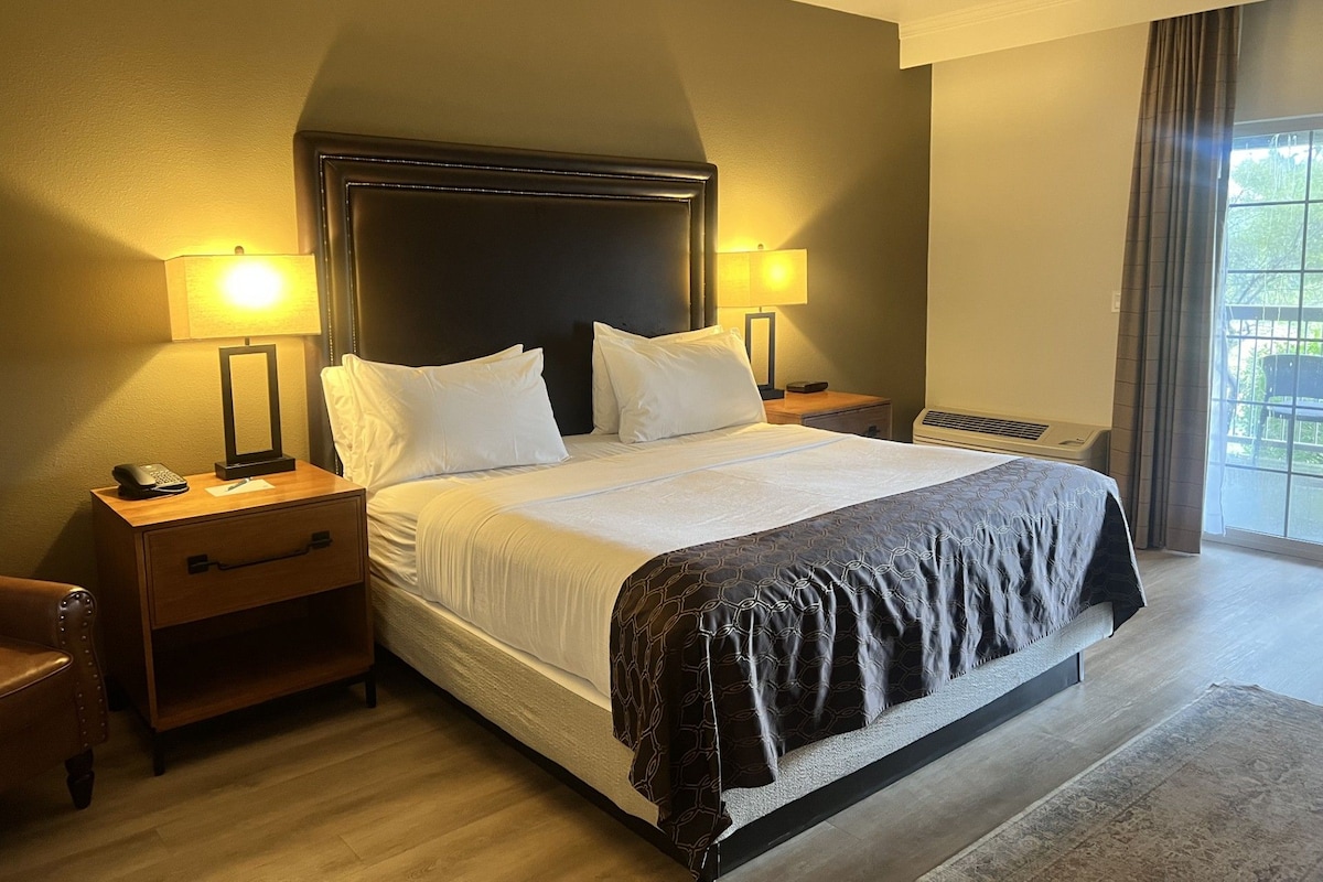 A hotel room with a large bed, two bedside tables with lamps, a wooden nightstand, a chair, and a window with curtains.