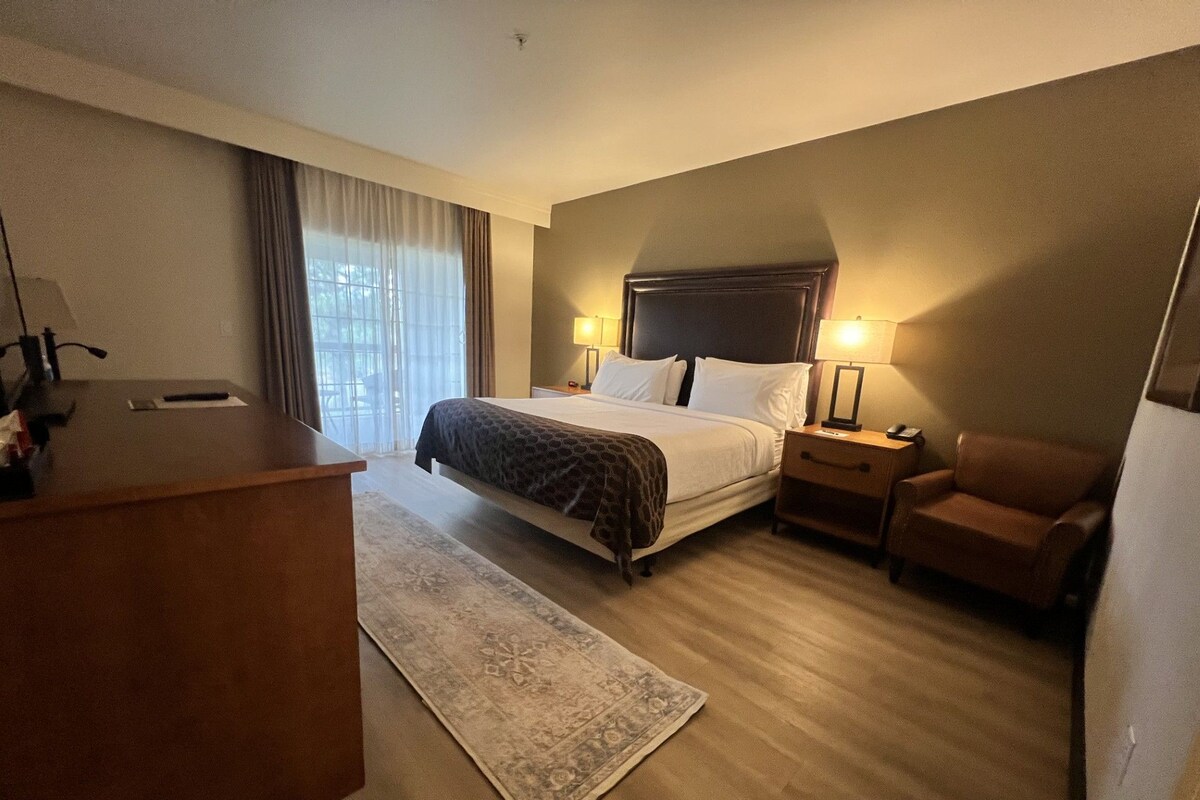A hotel room with a bed, bedside tables, a desk, a chair, and a large window with curtains.