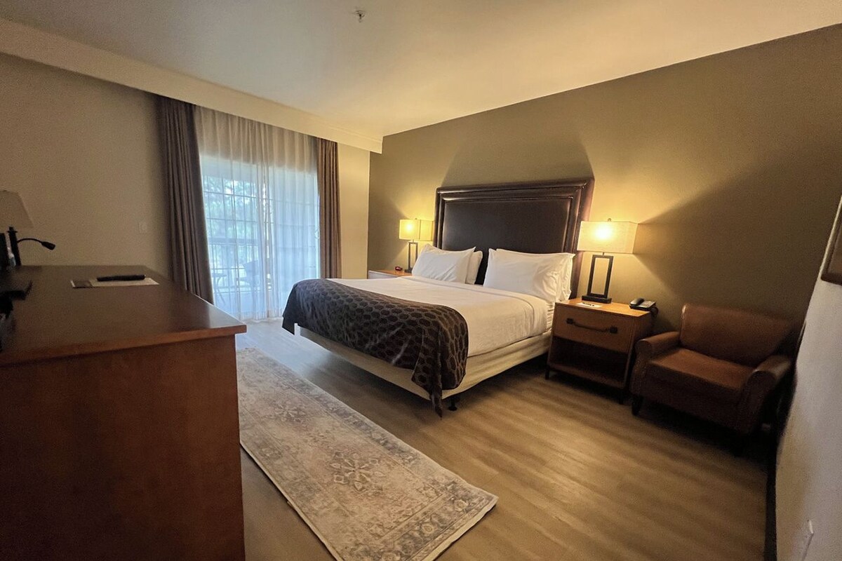 A hotel room with a bed, bedside tables, a desk, a chair, and a window with curtains.