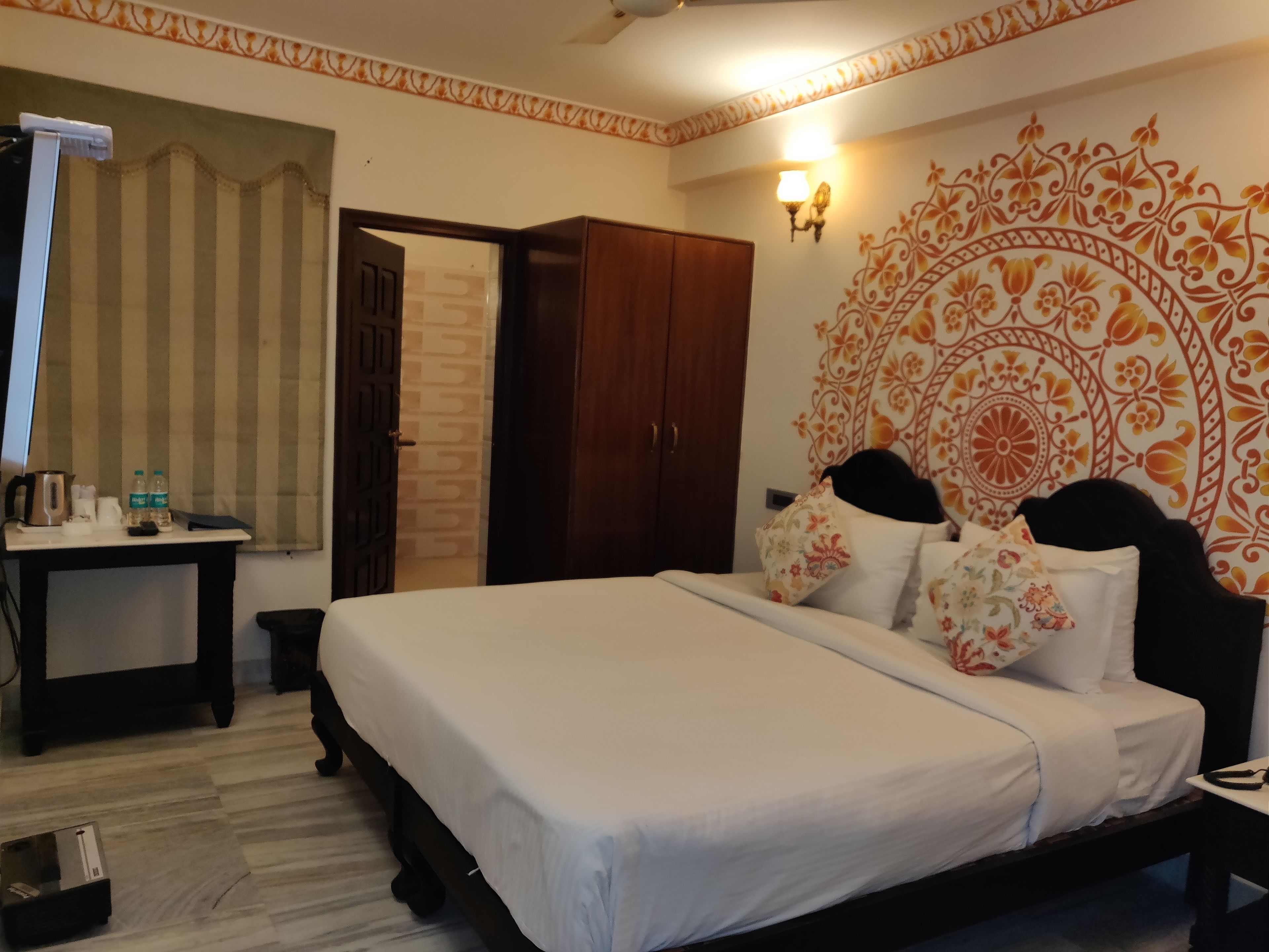 A hotel room with a bed, a desk with a kettle, two bottles of water, and a wall with a decorative pattern.