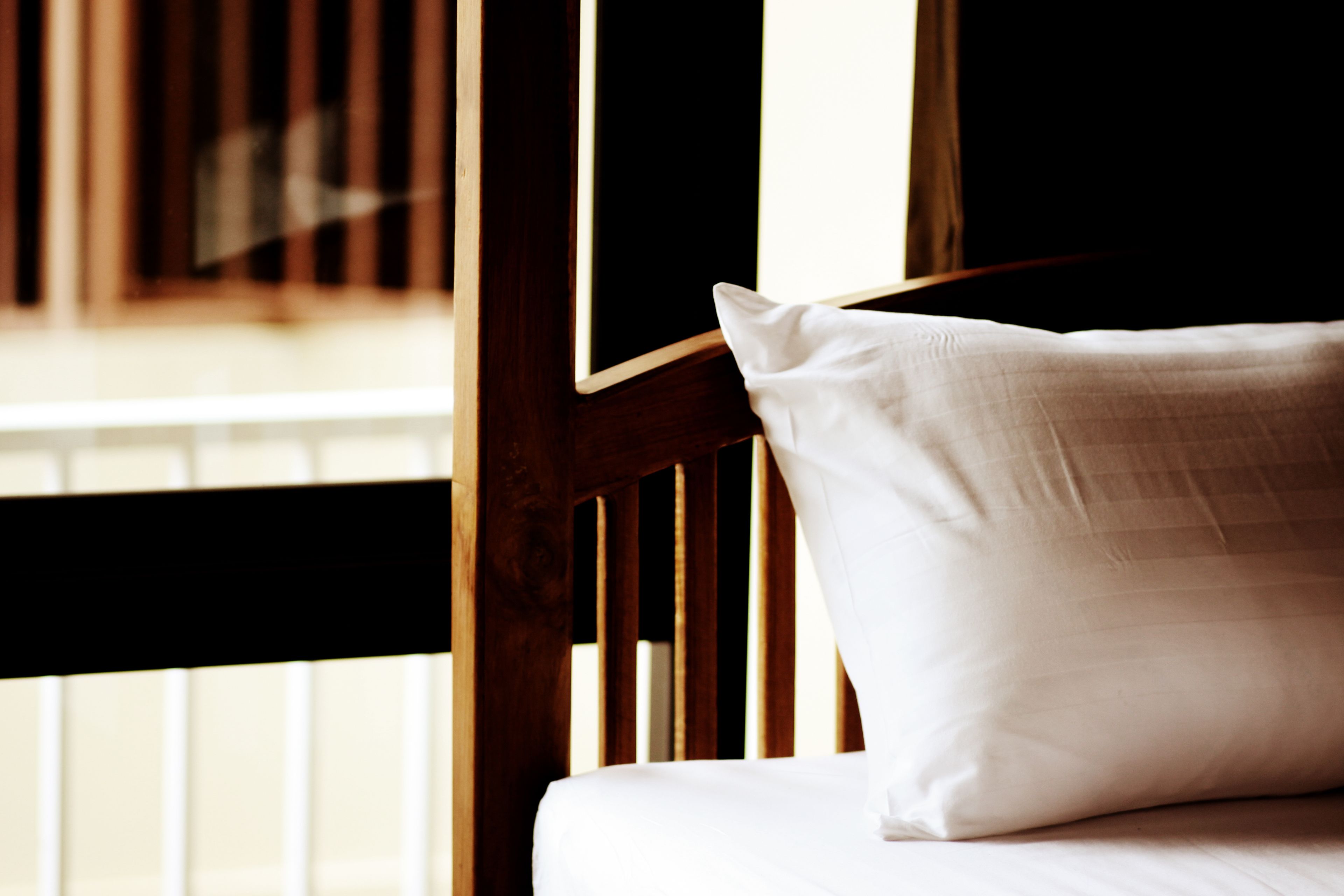 A bed with a white pillow and wooden headboard, positioned near a window with vertical blinds.