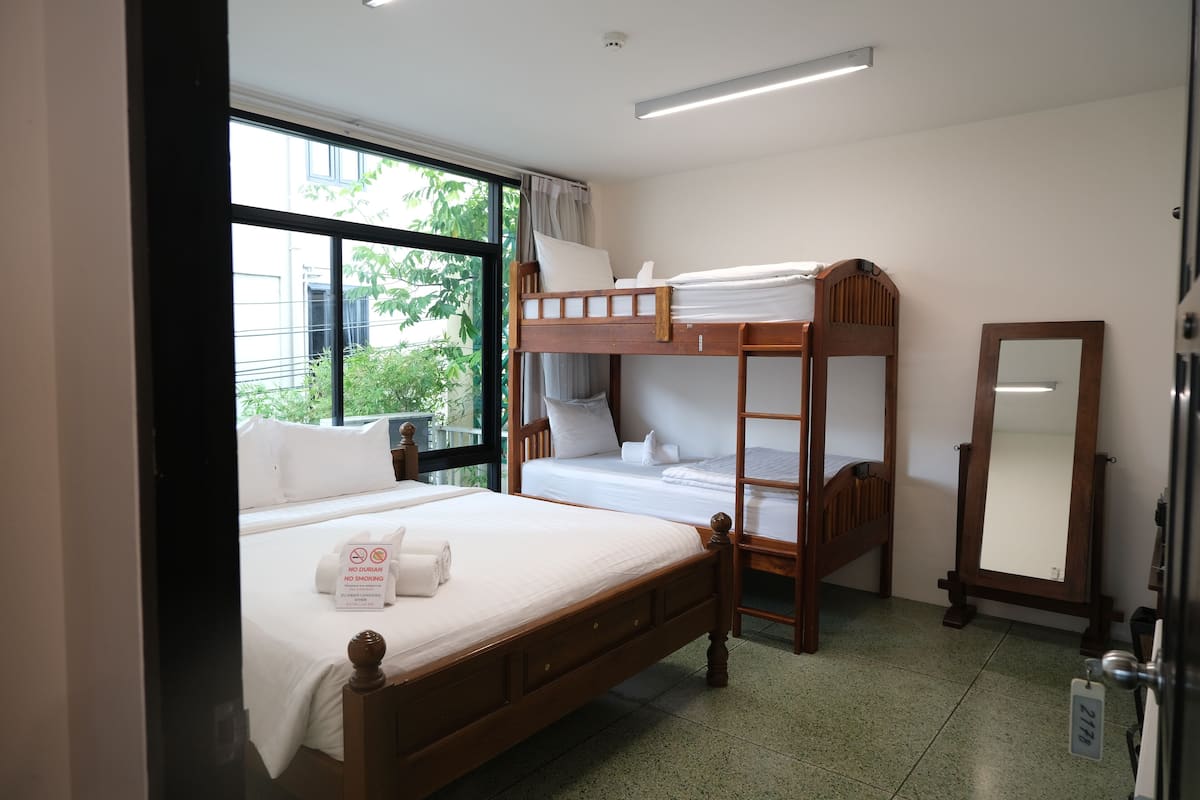 A room with a bunk bed, a single bed, a mirror, and a window with a view of greenery.