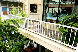 Un balcón con barandilla blanca, plantas en macetas y vistas a una habitación con una cama y una persona sentada.