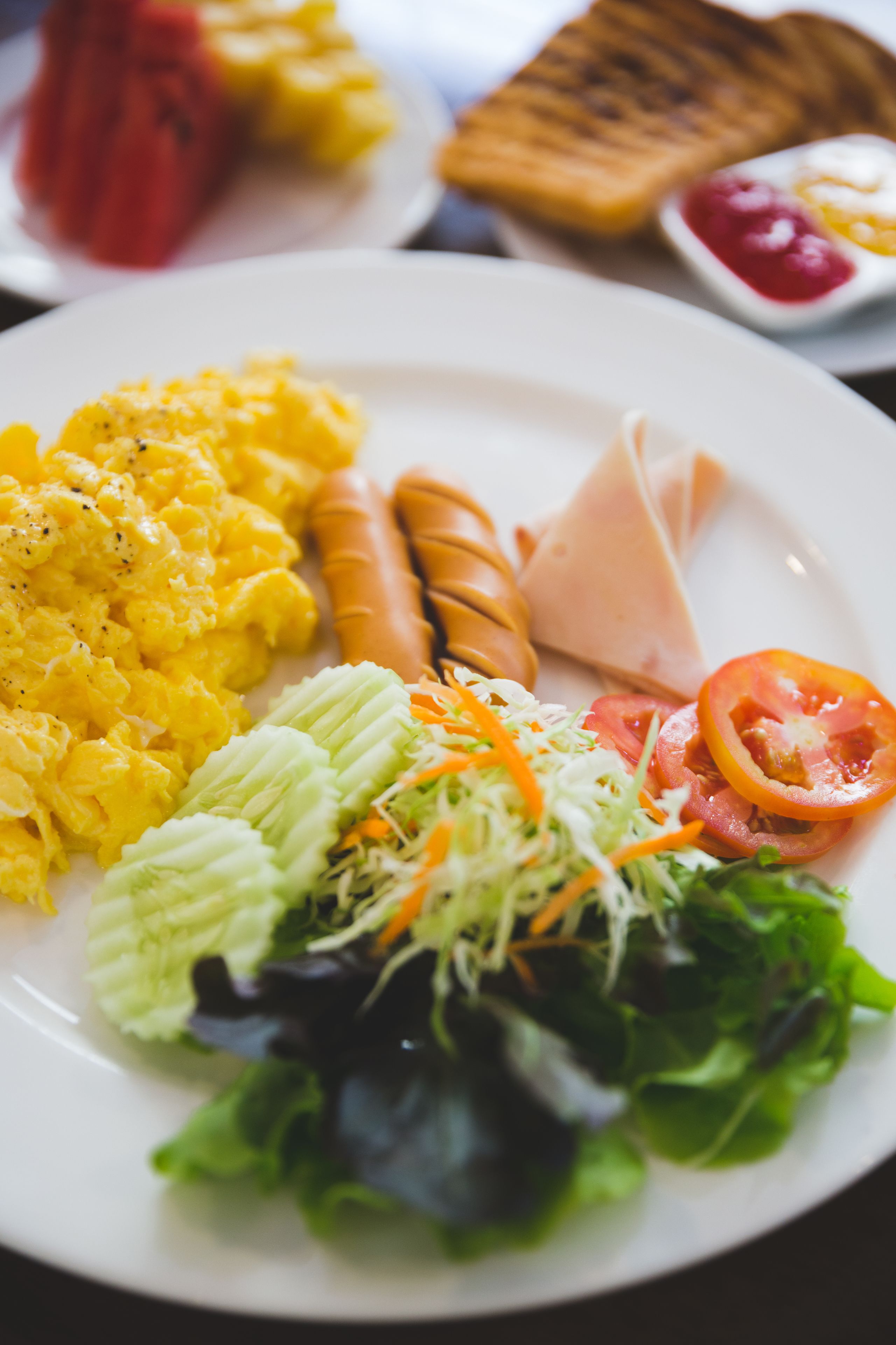 A plate of scrambled eggs, sausages, ham, and a salad with lettuce, tomatoes, and shredded carrots.