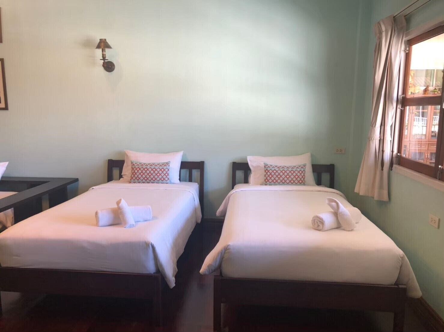 A room with two beds, each with a pillow and a rolled towel. There is a nightstand with a lamp between the beds.
