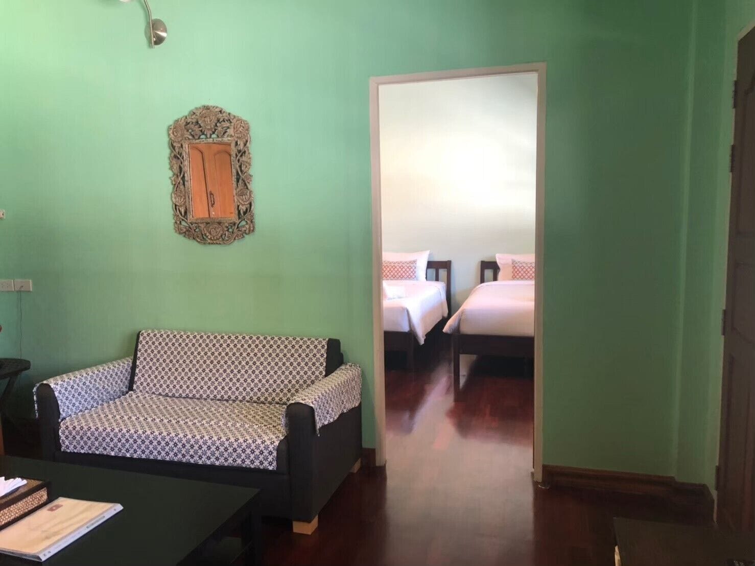 Une pièce avec un canapé à motifs, une petite table sur laquelle se trouve un classeur, un miroir et une porte qui donne sur une chambre à coucher avec deux lits.