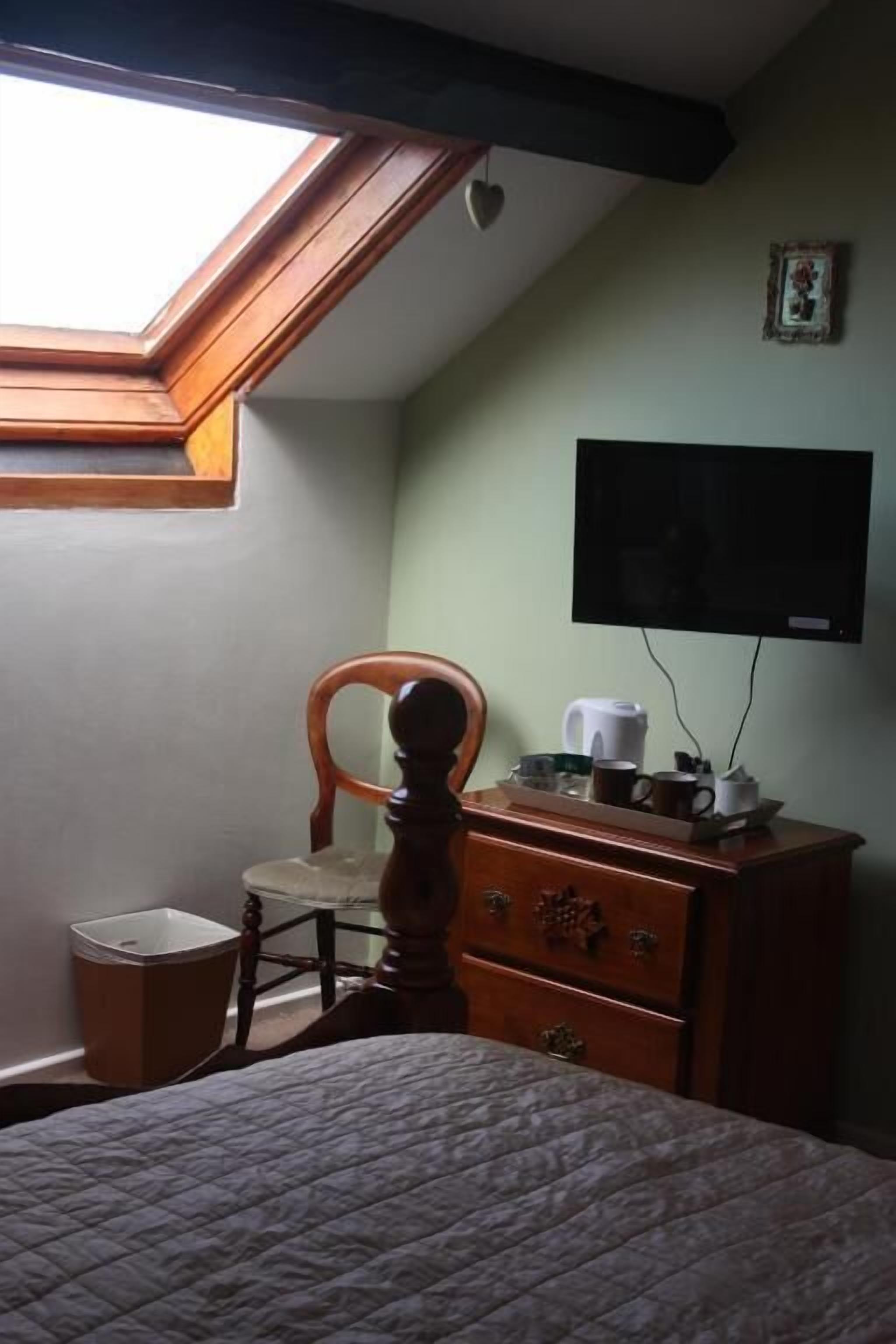 Un dormitorio con techo inclinado, un aparador de madera, una silla, una mesita de noche y un televisor montado en la pared.