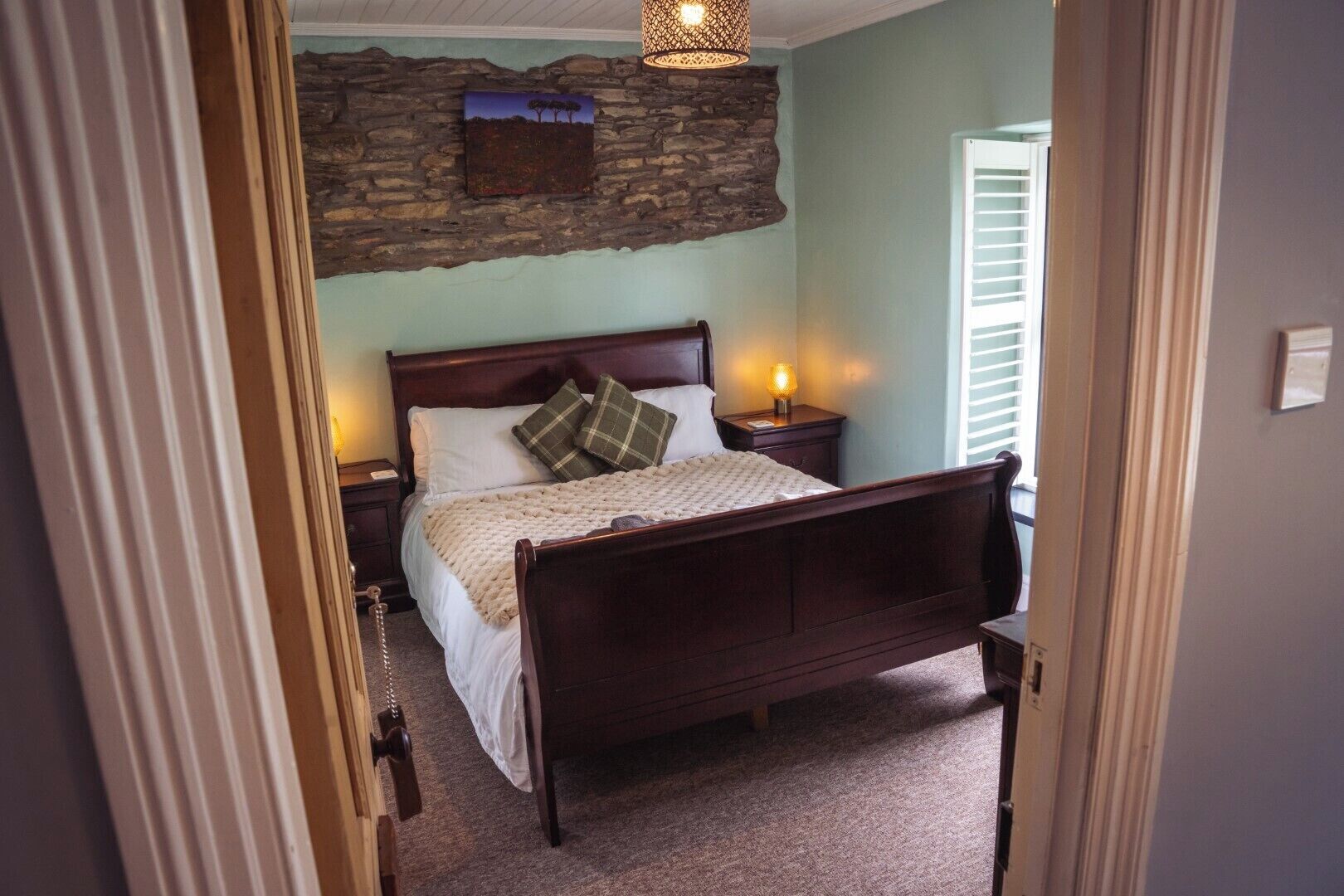 A bedroom with a stone wall, a wooden bed, a nightstand with a lamp, and a door leading to another room.