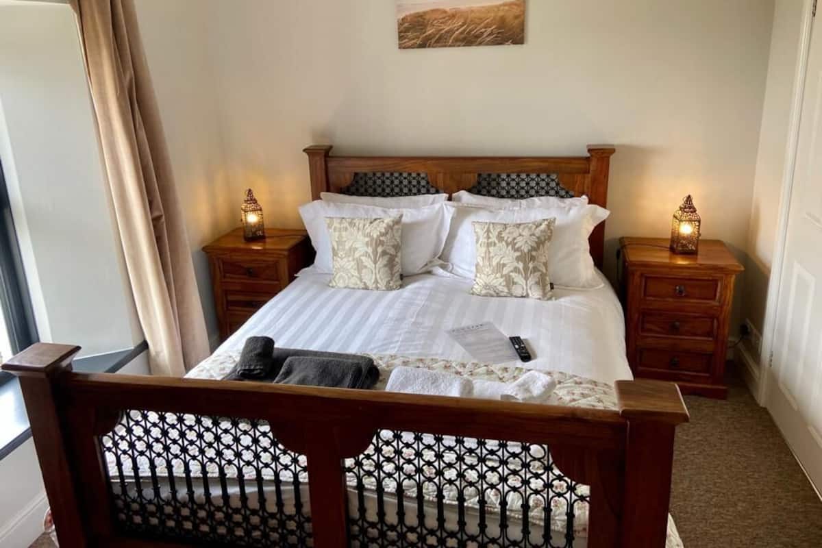 Un lit bien fait, avec du linge de lit blanc et des coussins à motifs, deux tables de chevet avec des lampes, une tête de lit en bois, une fenêtre avec des rideaux et un tableau accroché au mur.